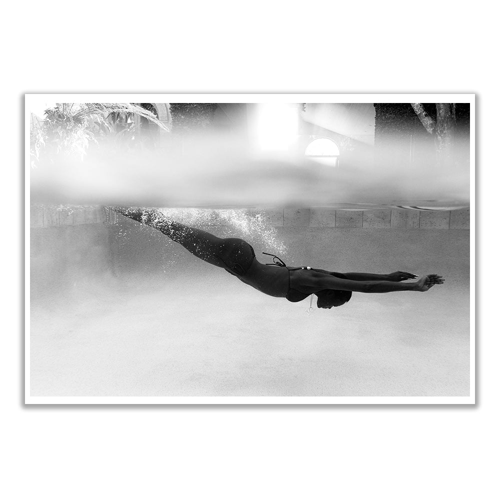 Black and white image of a person in a swimsuit diving underwater in a pool. Sunlight filters from above, creating a serene effect. The pool's surface is a distinct line across the middle.