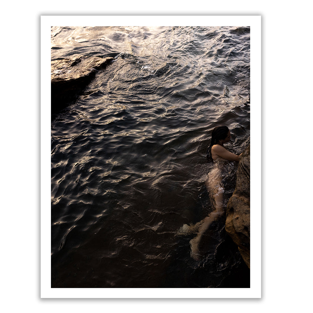 A person in a dark swimsuit is partly submerged in water, holding onto a rock formation. The water appears slightly turbulent, with reflections of the sky and surroundings visible on the surface. The scene is dimly lit, suggesting early morning or late afternoon—a moment captured as if painted with archival pigments in "Sunrise Swim #5" by Francesca Owen.