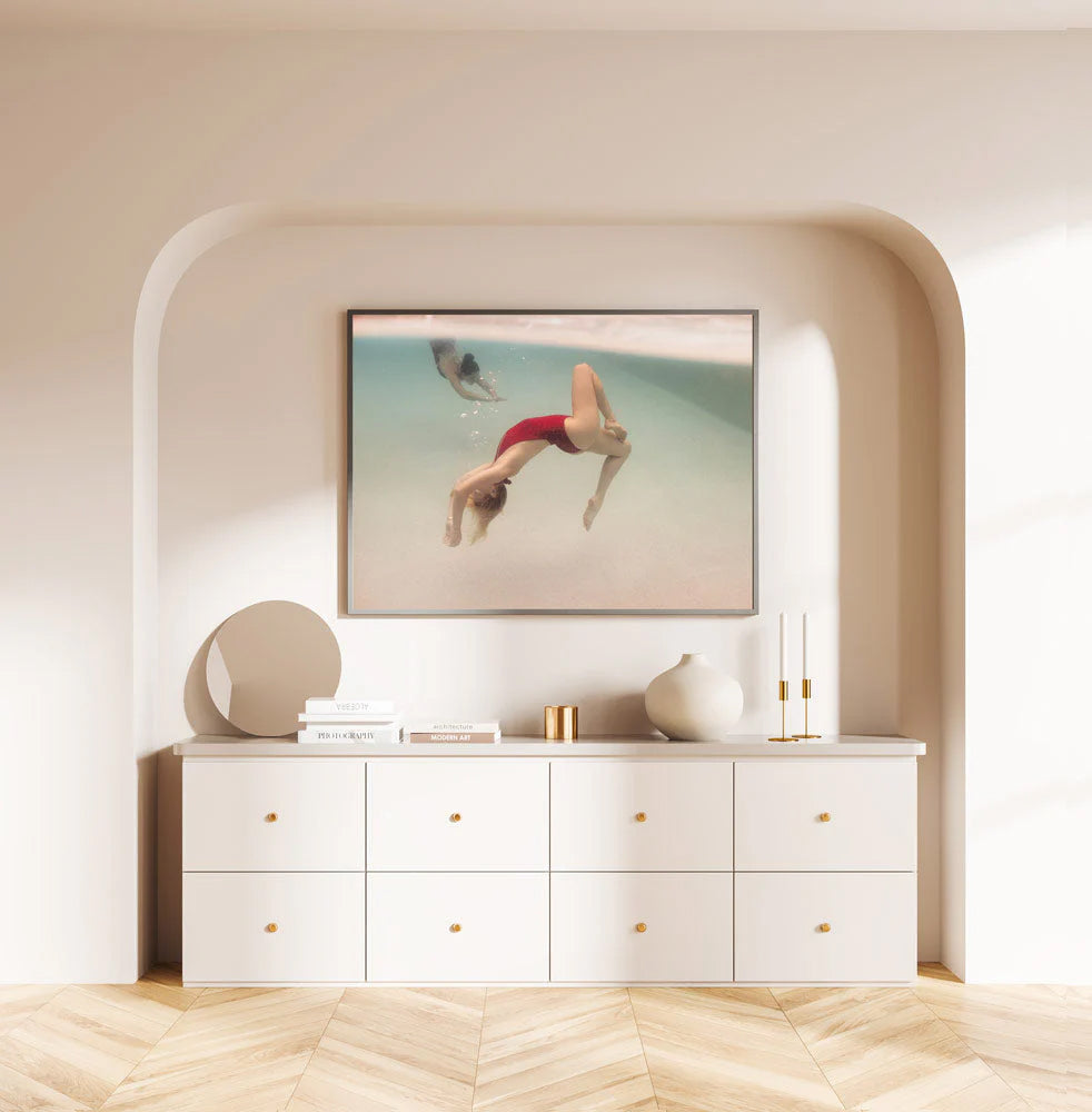 A minimalist room features a wooden floor and a white sideboard, with "Opposites Attract 01" by Francesca Owen hanging above it. The photograph captures a person swimming underwater, printed using archival pigments on fine art paper. The sideboard is adorned with round decor, creating a calm and serene atmosphere.