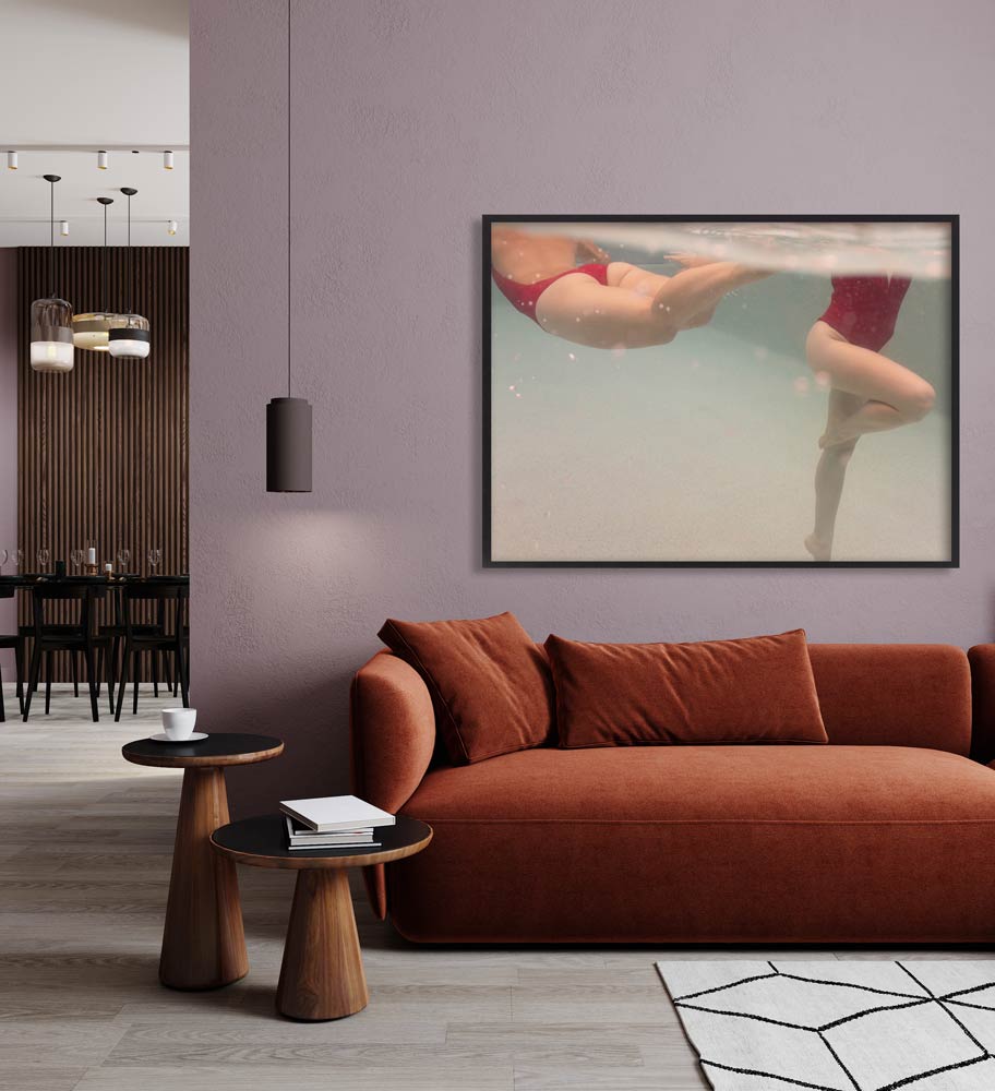 A modern living room features a rust-colored sofa with orange cushions, two wooden side tables, and a pendant light. On the wall is "Opposites Attract 03," a large underwater photograph of swimmers in red swimsuits by Francesca Owen, printed using archival pigments for longevity. In the background is a dining area with dark furniture and elegant lighting.