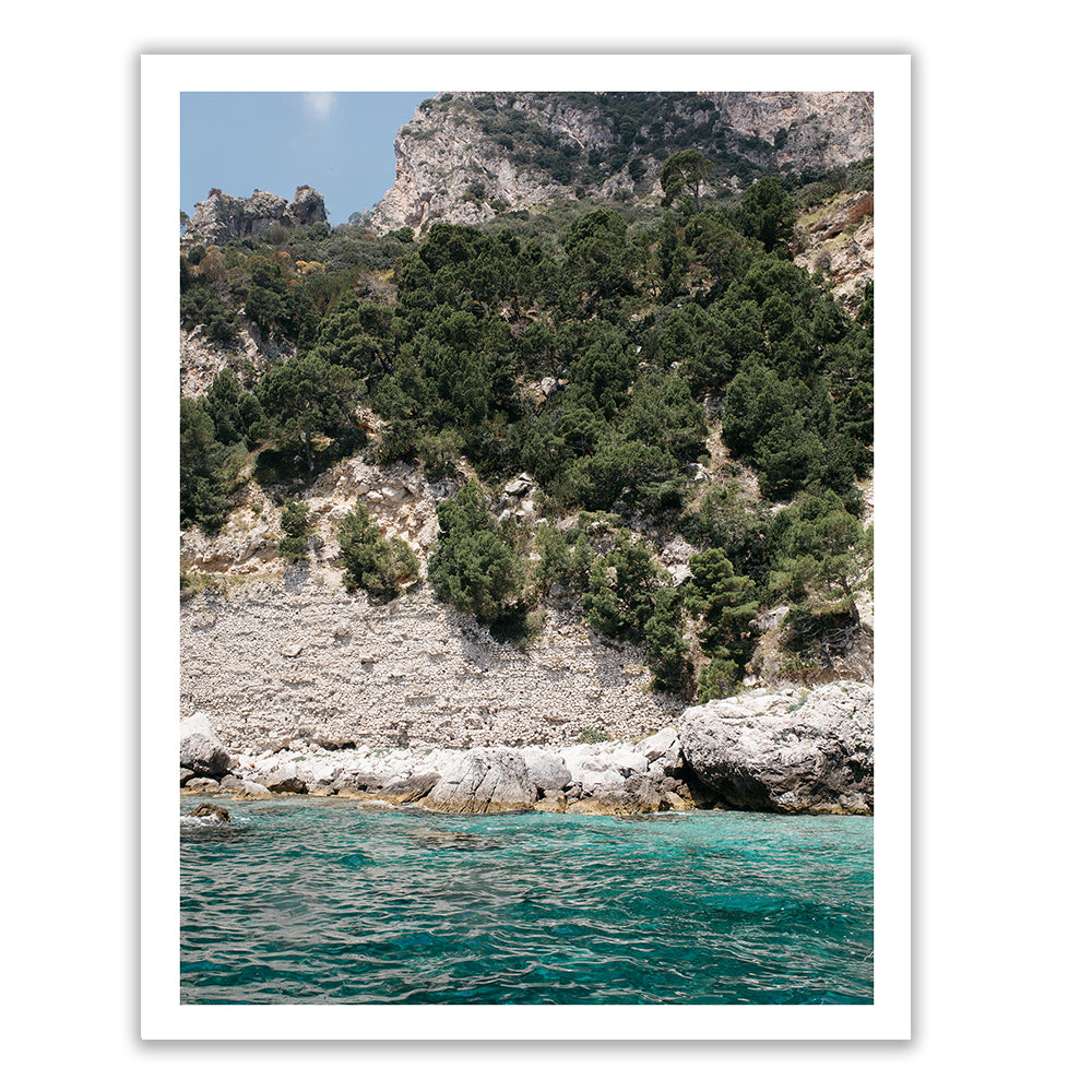 Aqua Waters #2 by Francesca Owen depicts a scenic coastal view of a rocky cliff covered with trees and greenery. The turquoise water at the base contrasts with the rugged shoreline, while the steep cliff rises sharply into the sky. This stunning image, crafted using archival pigments, is bordered by a white frame and comes with a certificate of authenticity.