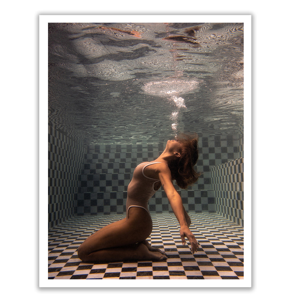 Underwater view of a person kneeling on a checkerboard-patterned pool floor, arching their back and exhaling bubbles towards the surface. The person's hair flows freely in the water as they wear a one-piece swimsuit. This scene from "The Banya #1" by Francesca Owen is perfect for fine art prints with custom framing, brought to life using archival pigments.