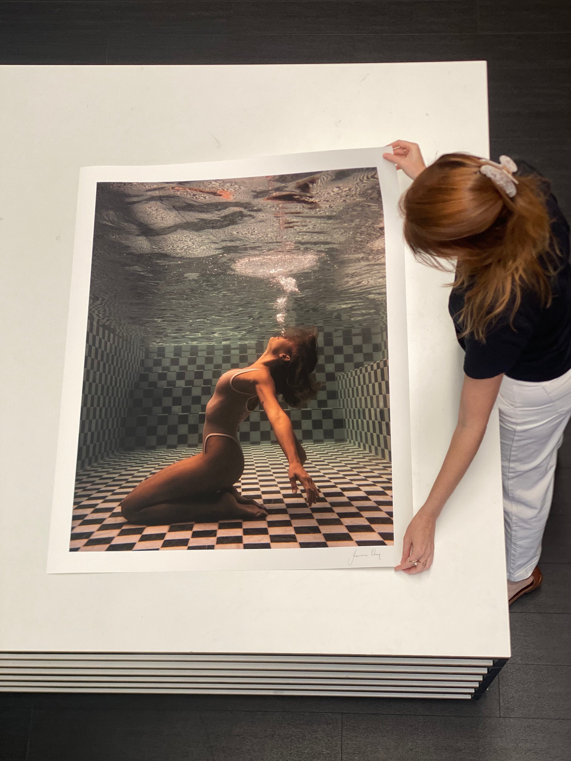 A woman with red hair secures the edges of a large, framed photograph. "The Banya #1," a fine art print created by Francesca Owen using archival pigments, depicts a person underwater in a pool, kneeling on a checkered black and white tiled floor, blowing bubbles towards the water's surface.