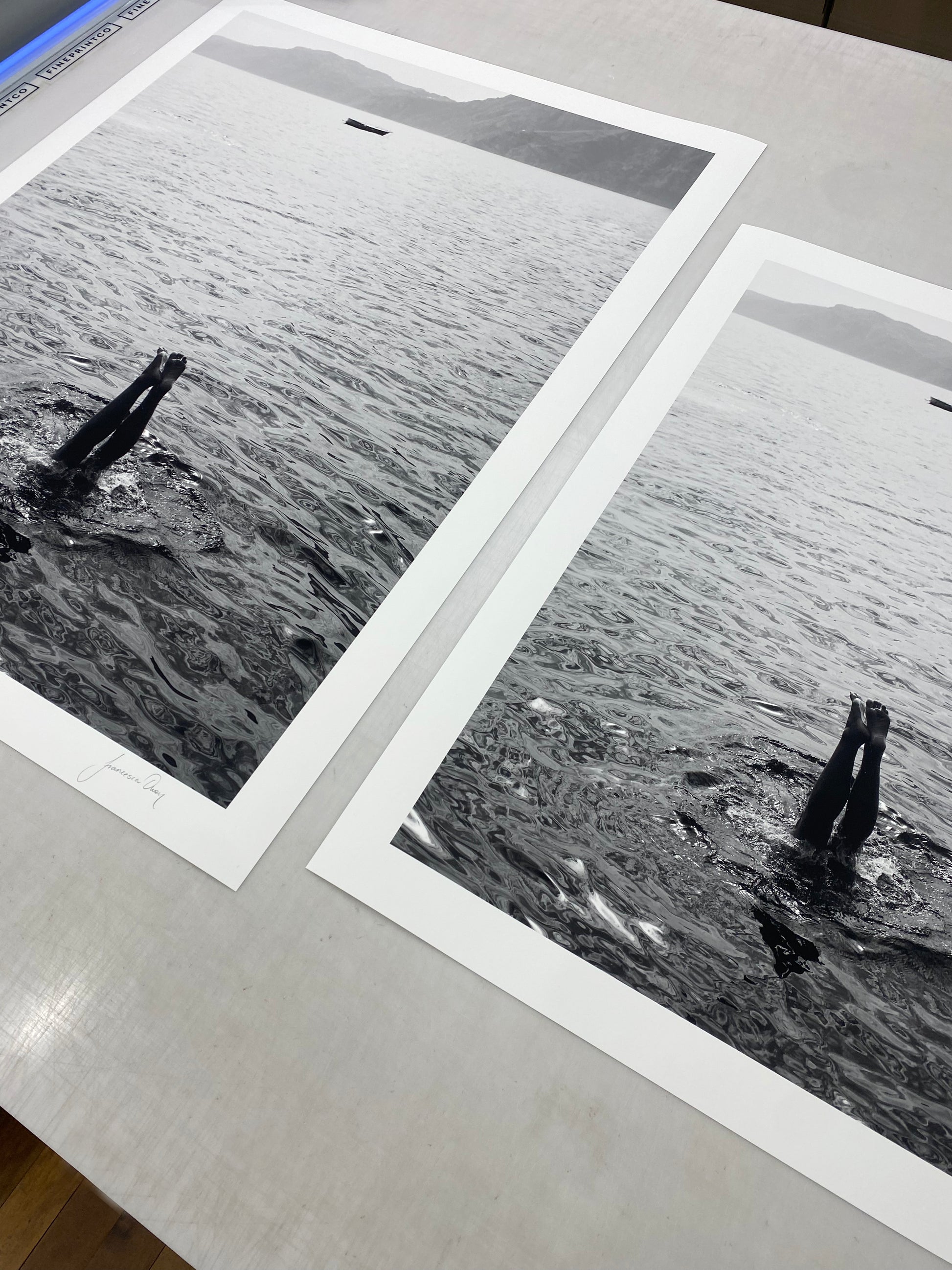 Capri Tippy Toes" by Francesca Owen captures a striking black and white image of a person performing a handstand in the water, with only their legs and feet visible above the surface. In the background, distant mountains and a solitary boat float on calm waters. This piece is available in custom sizes with archival pigments and includes a certificate of authenticity.