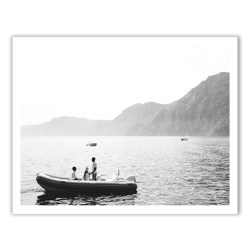 A black and white photo titled "Cruising," from the Francesca Owen collection, is printed with archival pigments on fine art paper and depicts three people standing on a small inflatable boat floating on a calm sea. Large rocky mountains and a few other distant boats are visible in the background of this serene scene, which is hand-signed by the artist.