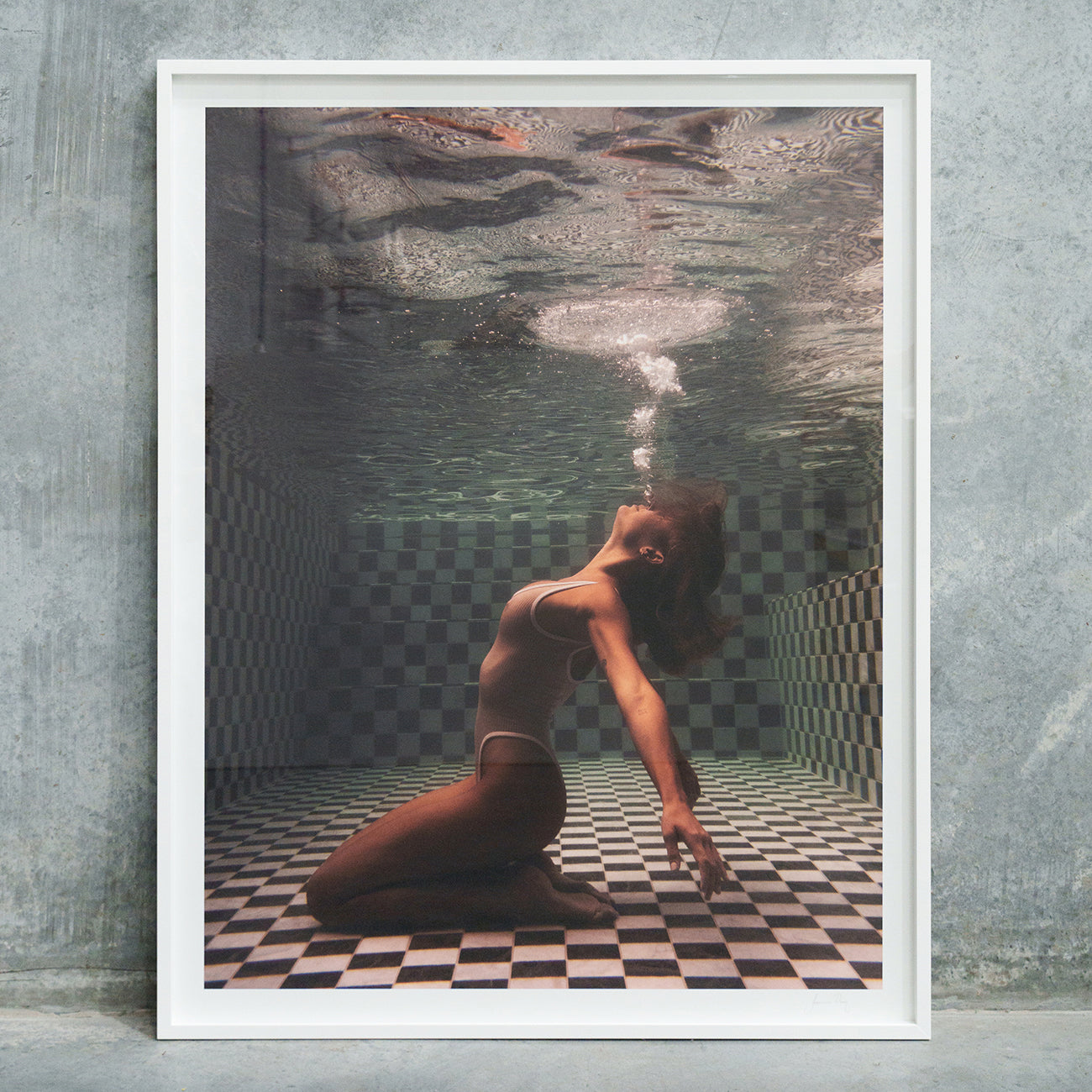 Francesca Owen's "The Banya #1" is a framed photograph depicting a person underwater, kneeling on a checkered floor, with their head tilted back and blowing bubbles upwards. Adorned in a one-piece swimsuit and seemingly submerged in a tiled pool, this striking image is created using archival pigments to ensure lasting quality and depth.