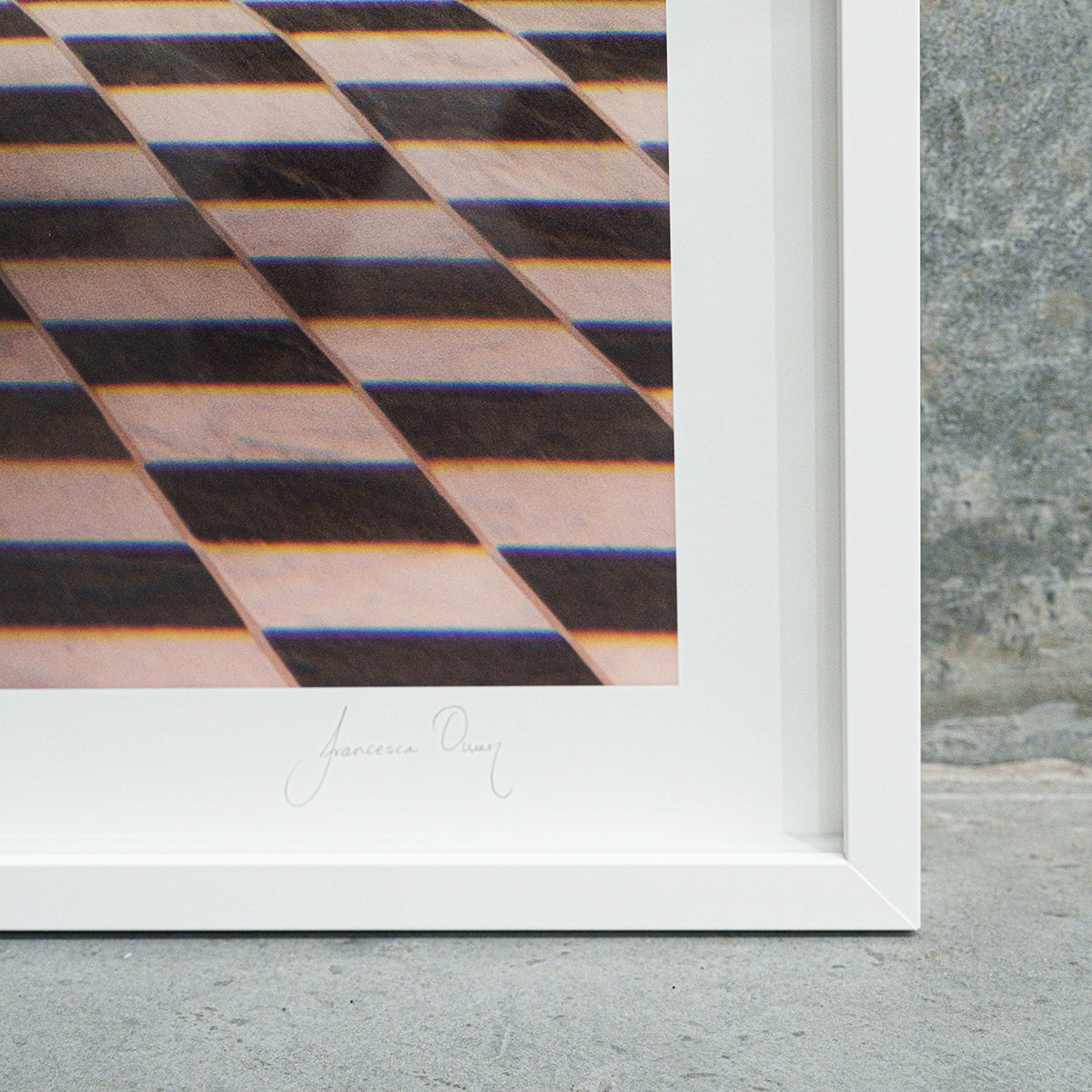 Product Description: "The Banya #1" by Francesca Owen is a framed photograph showcasing a checkerboard tile pattern with alternating light and dark tiles. It features a white frame and an image printed with archival pigments, signed in cursive near the bottom center. The backdrop behind the frame is a rough, grey concrete surface.