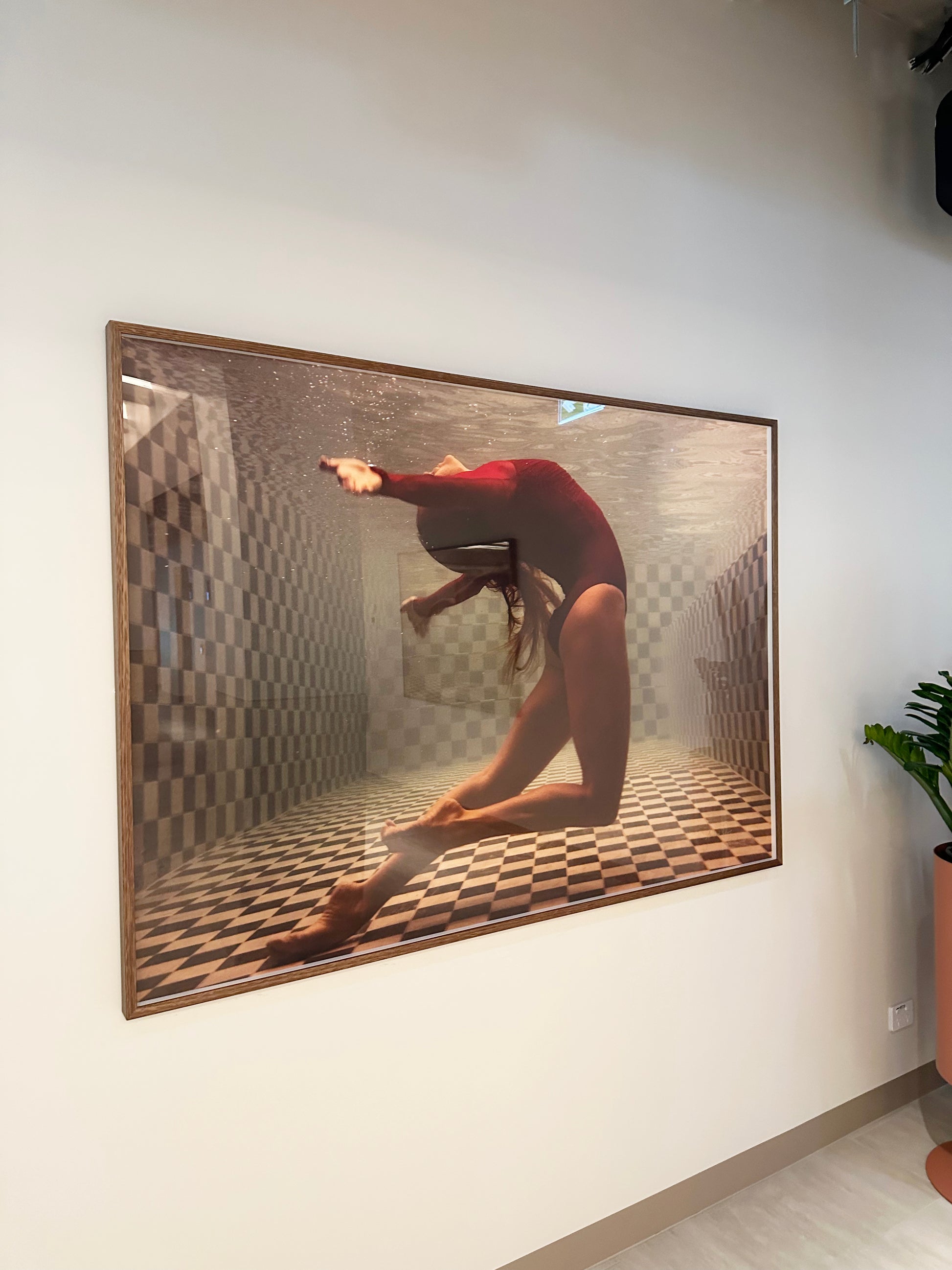 A photograph titled "Wild at Heart no.3" by Francesca Owen features a woman underwater in a red swimsuit, gracefully bending backwards. The pool setting includes a checkered pattern on the floor and walls, with archival pigments used for vibrancy. A potted plant is partially visible in the corner of this fine art piece.