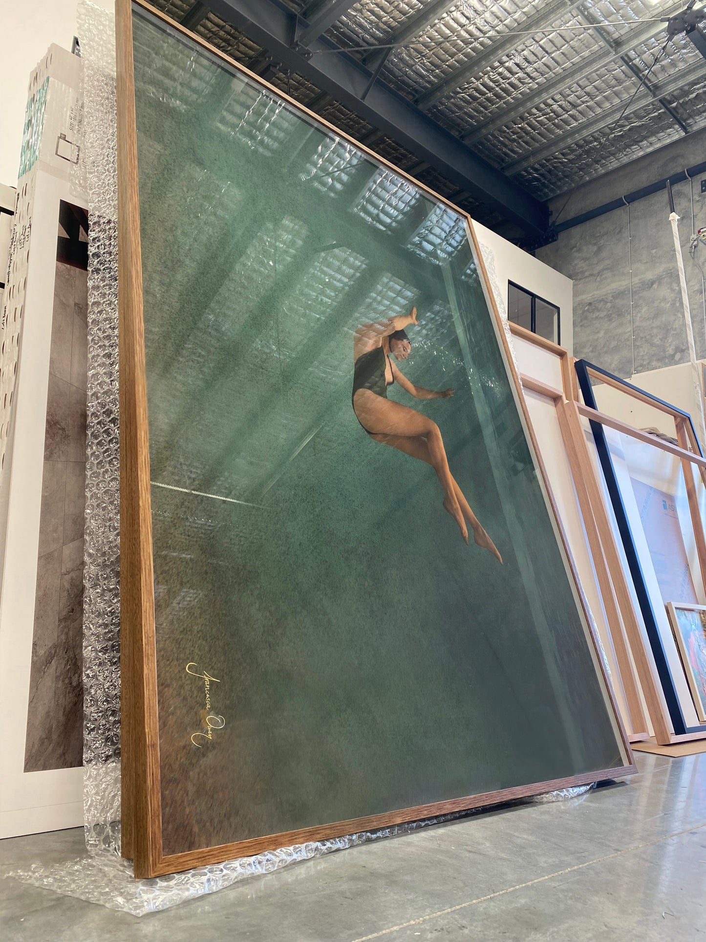 Silent Surrender #1, a large framed photograph from Francesca Owen, printed on fine art paper with archival pigments, leans against a wall in an art gallery or storage area. The image depicts a woman in a black swimsuit gracefully swimming underwater with light filtering through the surface. Other framed artworks are visible in the background.