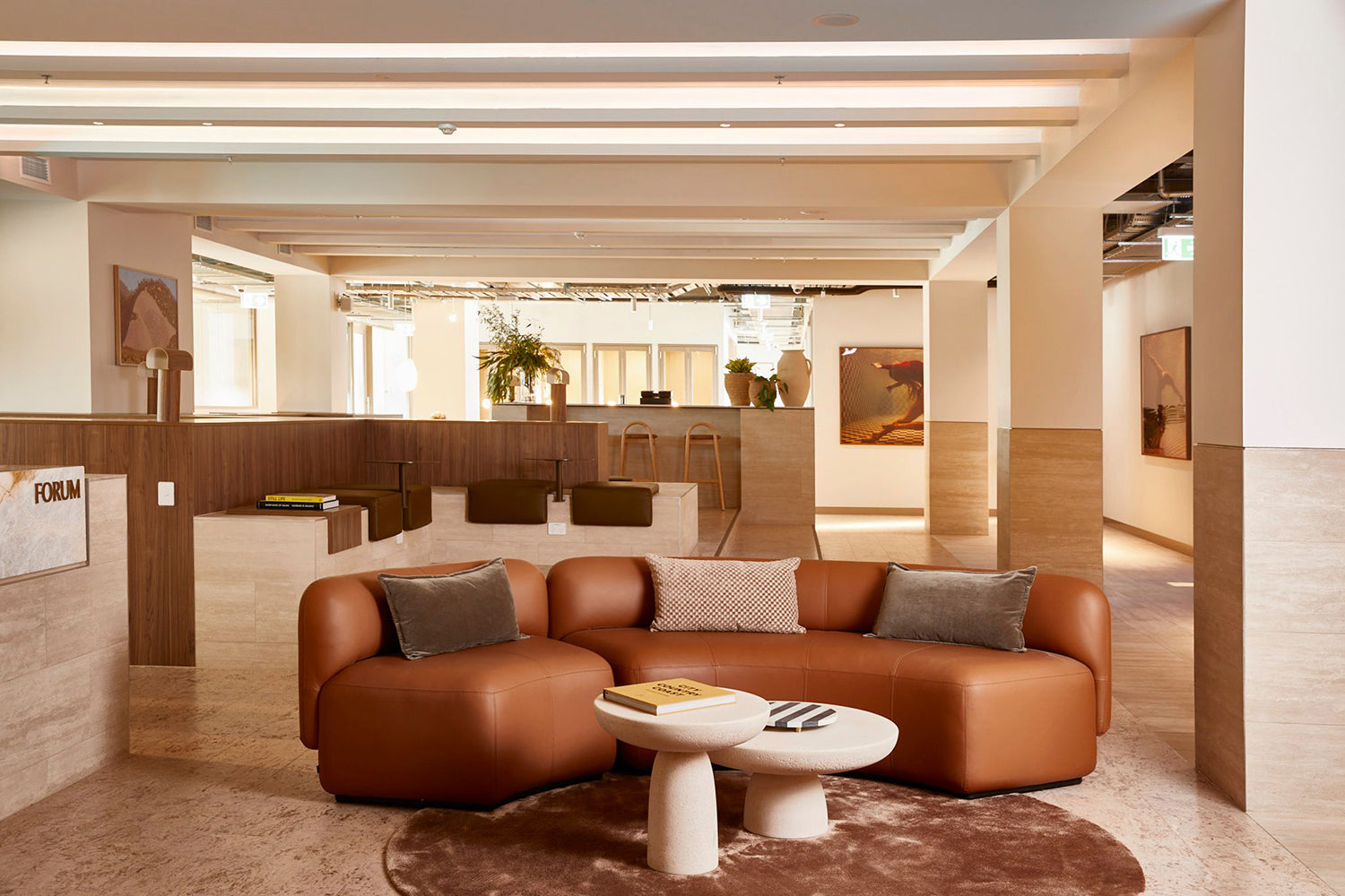 A modern lounge area featuring a custom-made curved brown leather sofa, a round coffee table, and decorative pillows. The space is well-lit, highlighting neutral tones and framed artwork titled "Wild at Heart no.3" by Francesca Owen on fine art paper. Additional seating enhances the elegant design.