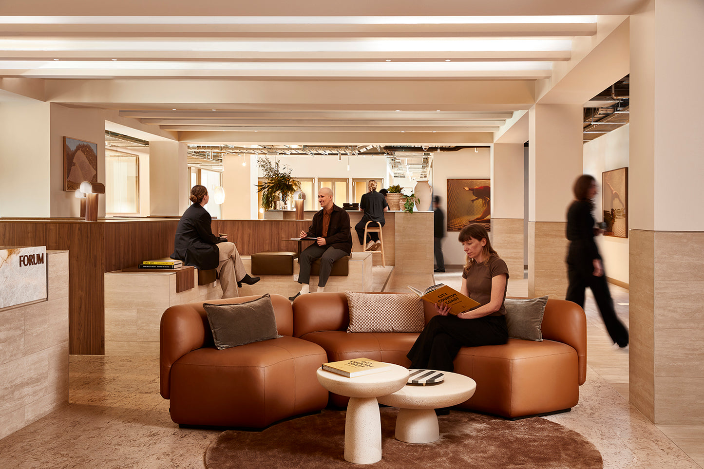 A modern, open office space boasts a custom-made brown leather couch with people seated and reading. Meanwhile, others engage in conversation at tables, and the walls are beautifully adorned with fine art paper prints from Francesca Owen's "Wild at Heart no.4" collection. Soft lighting and neutral tones create a welcoming atmosphere.