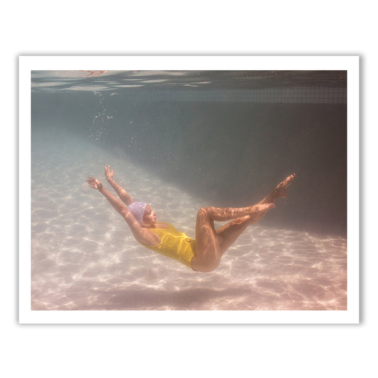 A person wearing a yellow swimsuit and a swim cap is captured underwater performing a graceful, floating pose. They are submerged in a well-lit swimming pool, with sunrays filtering through the water. This ethereal scene would make for stunning fine art paper prints using archival pigments as part of Francesca Owen's "Halcyon Daze Part 1.