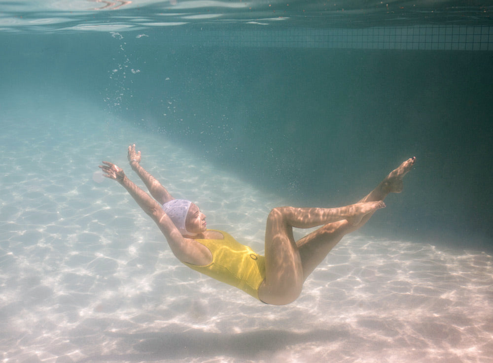 A person in a yellow swimsuit is swimming underwater in a pool, with their arms and legs extended gracefully. They are wearing a swim cap and appear to be performing a fluid, dance-like movement. The lighting creates a serene, clear atmosphere, perfectly captured as "Halcyon Daze 1" by Francesca Owen on fine art paper with archival pigments.