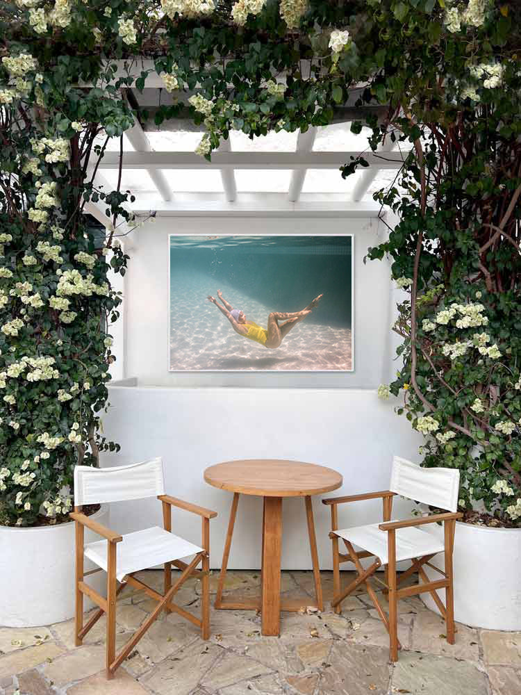 A cozy outdoor seating area features two white chairs and a wooden table between them, framed by lush greenery and climbing plants. On the wall behind the table hangs "Halcyon Daze 1," a fine art print of a person swimming underwater created with archival pigments by Francesca Owen, adding a serene touch to the scene.