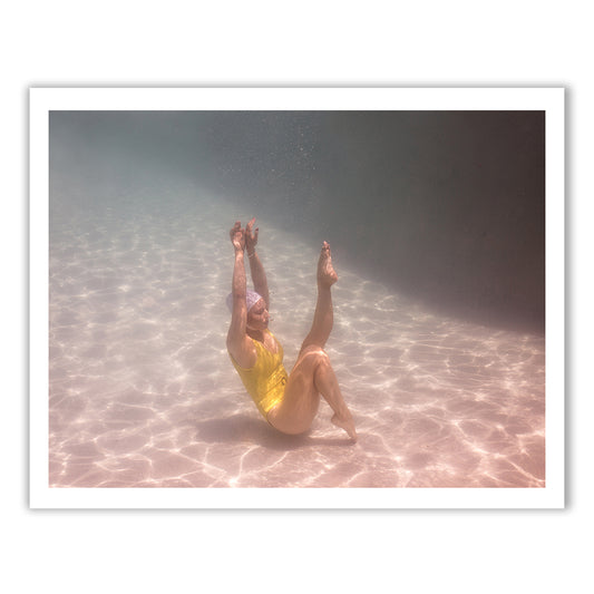 A person clad in a vibrant yellow swimsuit and swim cap glides effortlessly underwater in a pool, striking an elegant pose with arms and legs extended upwards. The sunlight dances through the water, creating intricate rippling patterns on the pool floor, evocative of Francesca Owen's "Halcyon Daze Part 3," where archival pigments craft exquisite artwork on fine art paper.