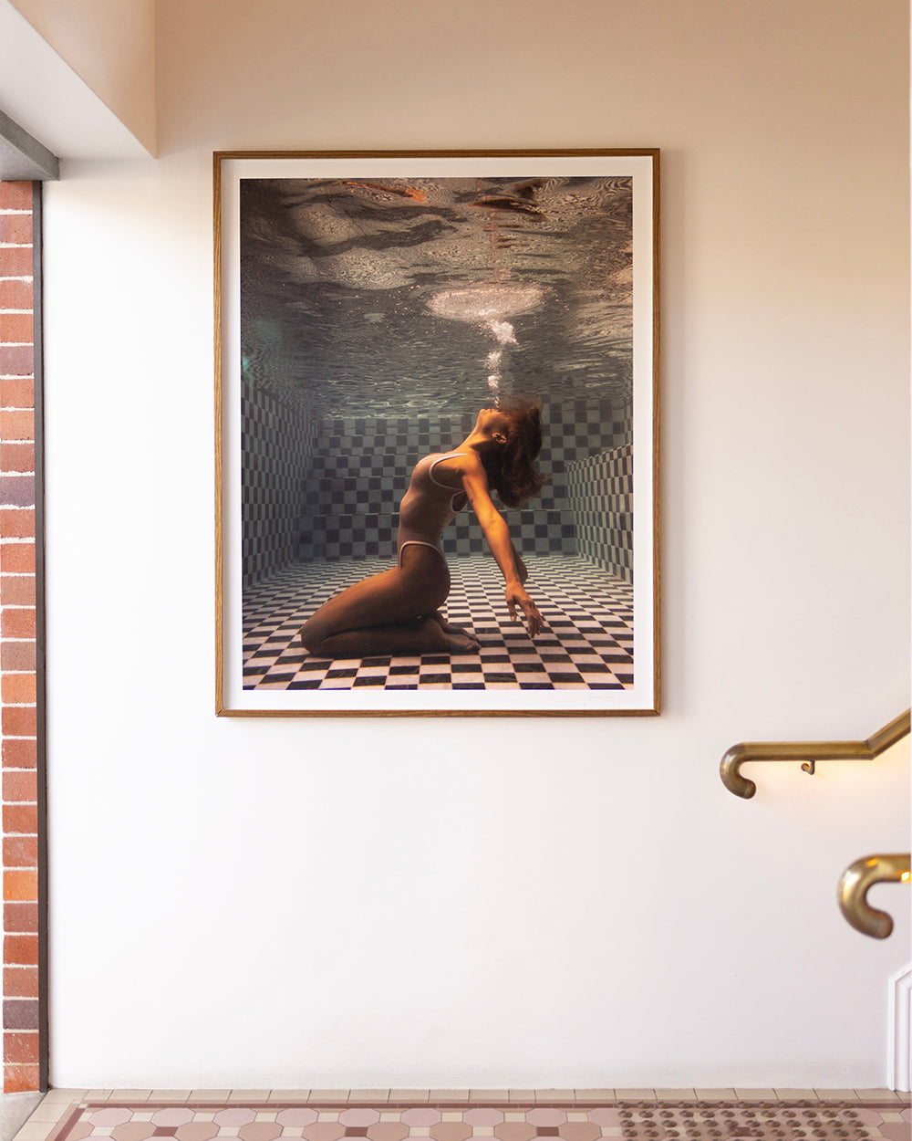 A framed photograph titled "The Banya #1" by Francesca Owen hangs on a light-colored wall beside a large window with a brick frame. The image, created with archival pigments, depicts a woman underwater, kneeling on a tiled pool floor, arching her back and exhaling a bubble of air. Another wall and a staircase railing are partially visible.