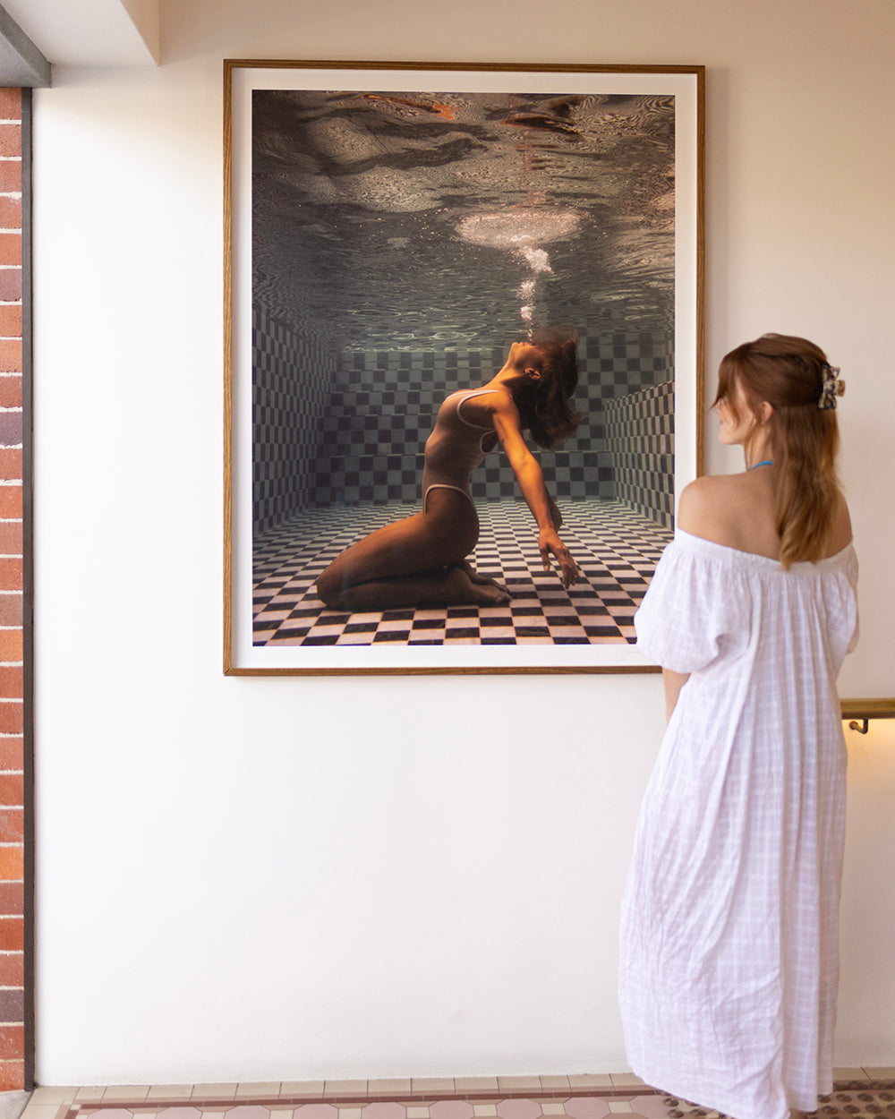 A person in a white off-shoulder dress stands in a gallery, observing "The Banya #1," a large framed photograph by Francesca Owen. The artwork, printed on fine art paper, depicts someone underwater, kneeling on checkerboard tiles while exhaling bubbles towards the surface.