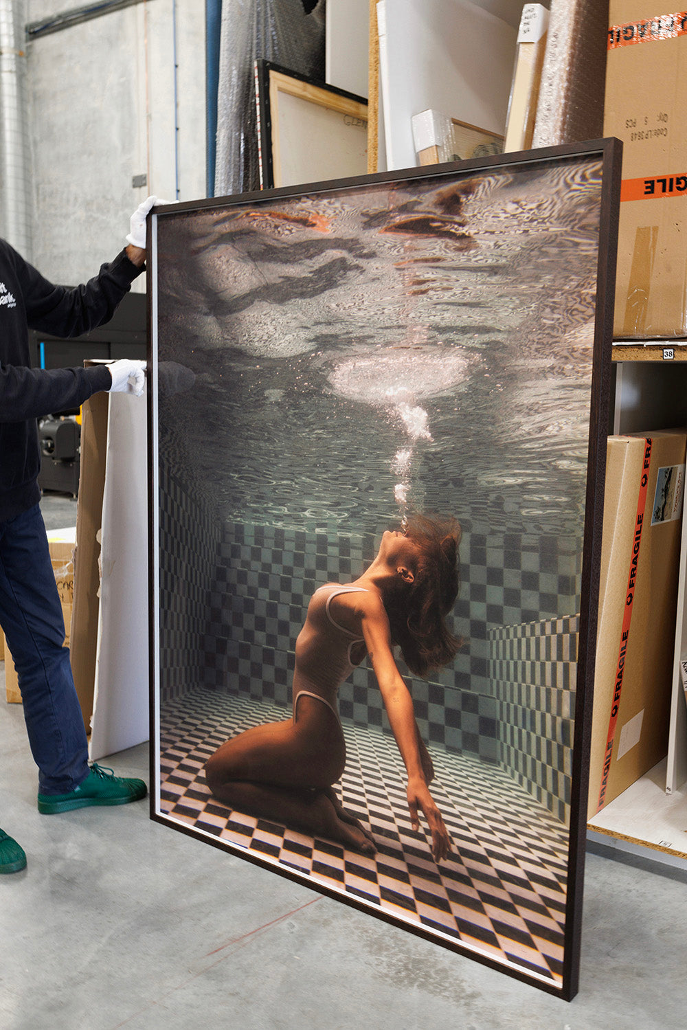 A person wearing gloves holds up a large framed photograph. "The Banya #1" by Francesca Owen depicts a woman underwater, arching her back and blowing bubbles. She is in a checkered pool, wearing a bikini. The background shows various cardboard boxes and packing materials, suggesting custom framing with archival pigments.