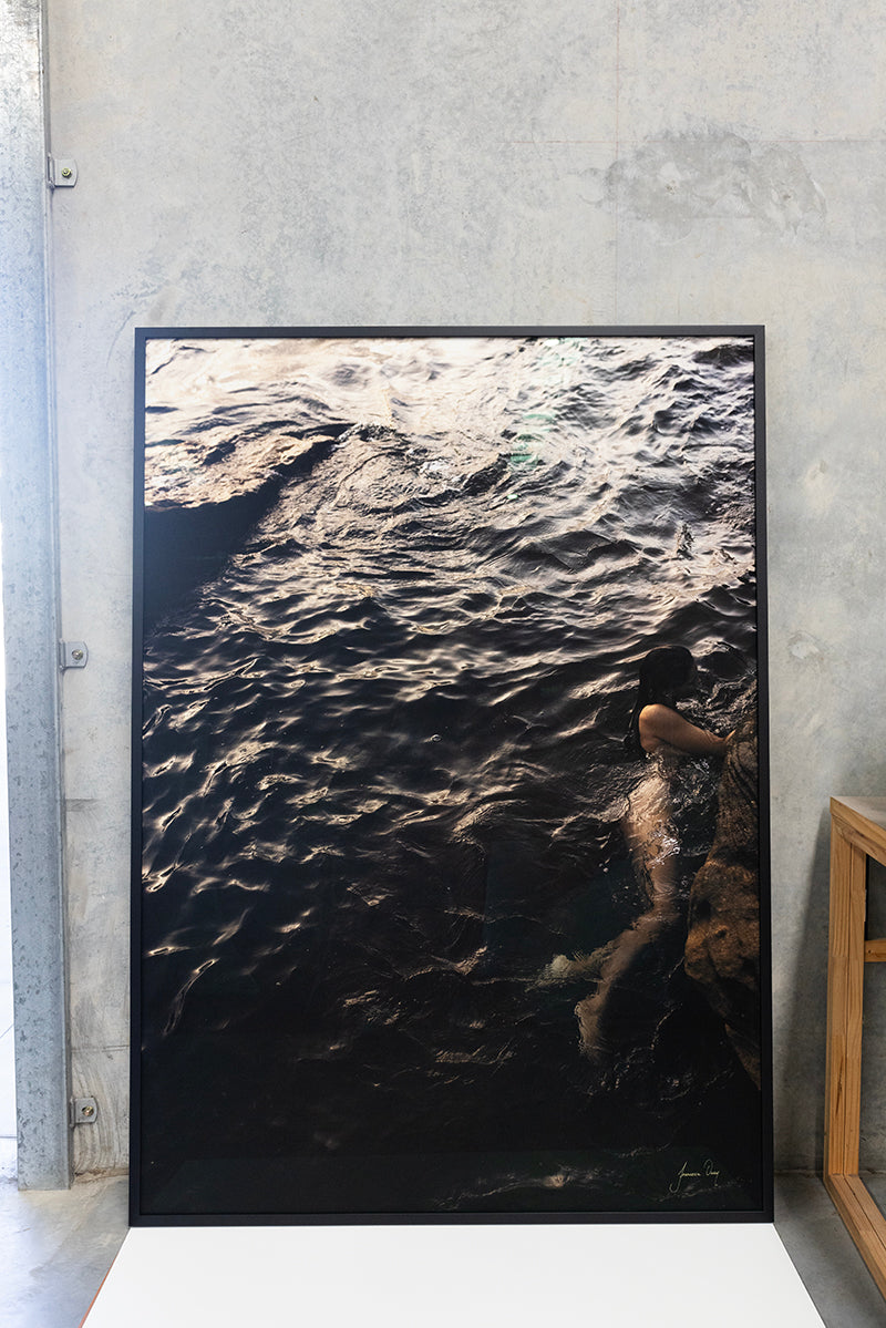 A framed photograph titled "Sunrise Swim #5" by Francesca Owen depicts a person submerged in dark, swirling water, propped up against a concrete wall. The moody lighting emphasizes the texture of the water. Printed with archival pigments on fine art paper, it also includes a certificate of authenticity.