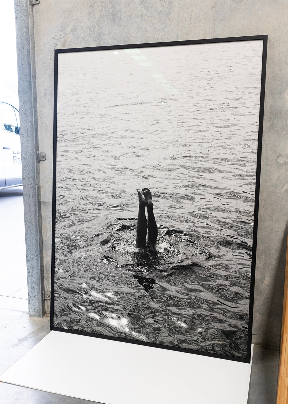 Capri Tippy Toes" by Francesca Owen captures a striking black and white image of a person performing a handstand in the water, with only their legs and feet visible above the surface. In the background, distant mountains and a solitary boat float on calm waters. This piece is available in custom sizes with archival pigments and includes a certificate of authenticity.