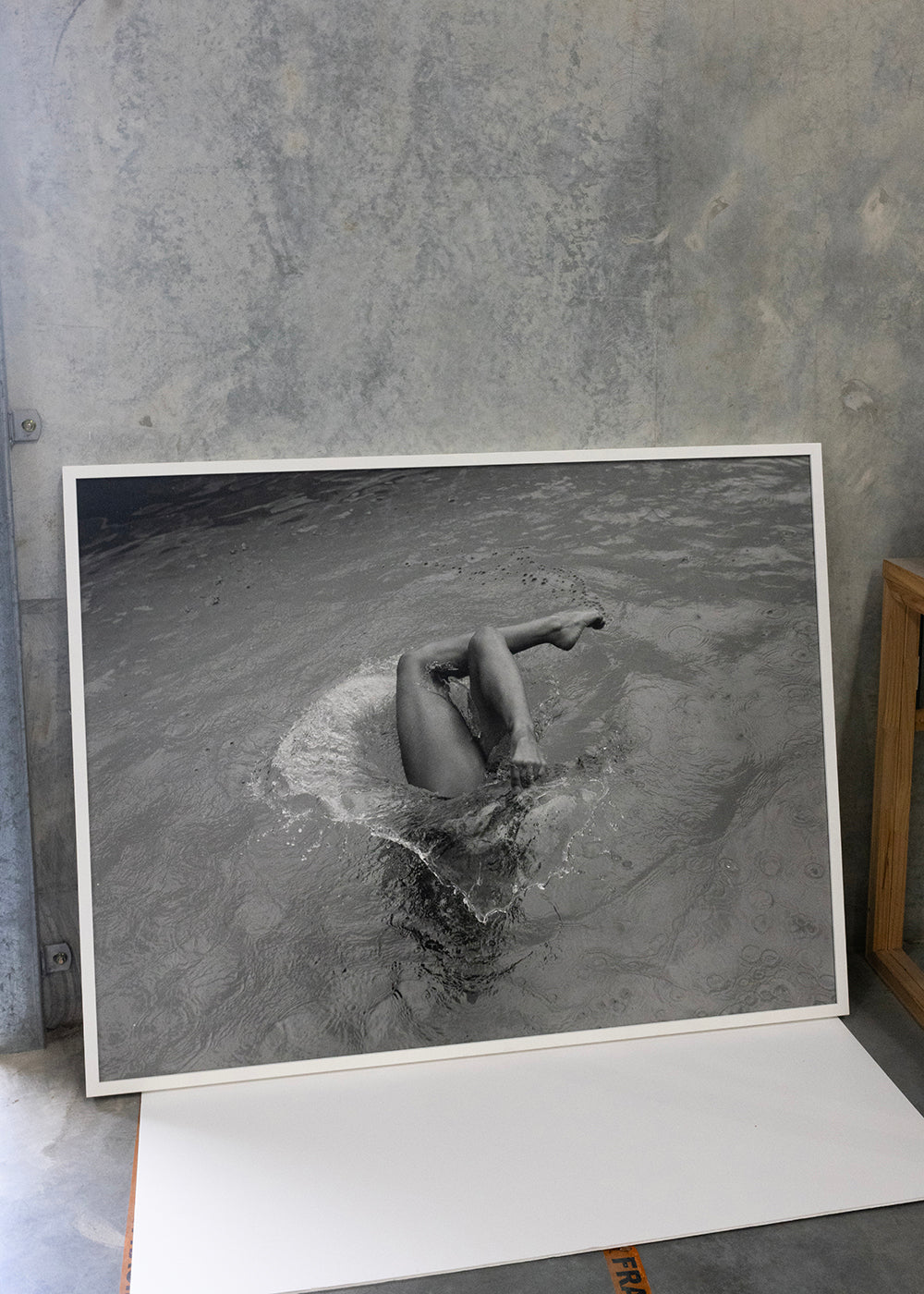 Self Reflection," a fine art print by Francesca Owen created with archival pigments, features a black-and-white photograph of a person partially submerged in water, with their legs and feet visible above the surface as if they are mid-dive or floating. The framed artwork is propped against a concrete wall near a wooden shelf and scattered papers.