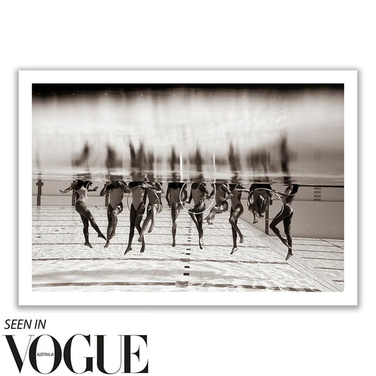 The artwork "Ladies in Waiting #3" by Francesca Owen features a black and white photograph of synchronized swimmers underwater in a pool, showcasing their elegantly extended limbs. This fine art piece highlights the swimmers' grace and coordination and is printed on limited edition giclee fine art paper. A note in the corner reads "Seen in Vogue Australia.