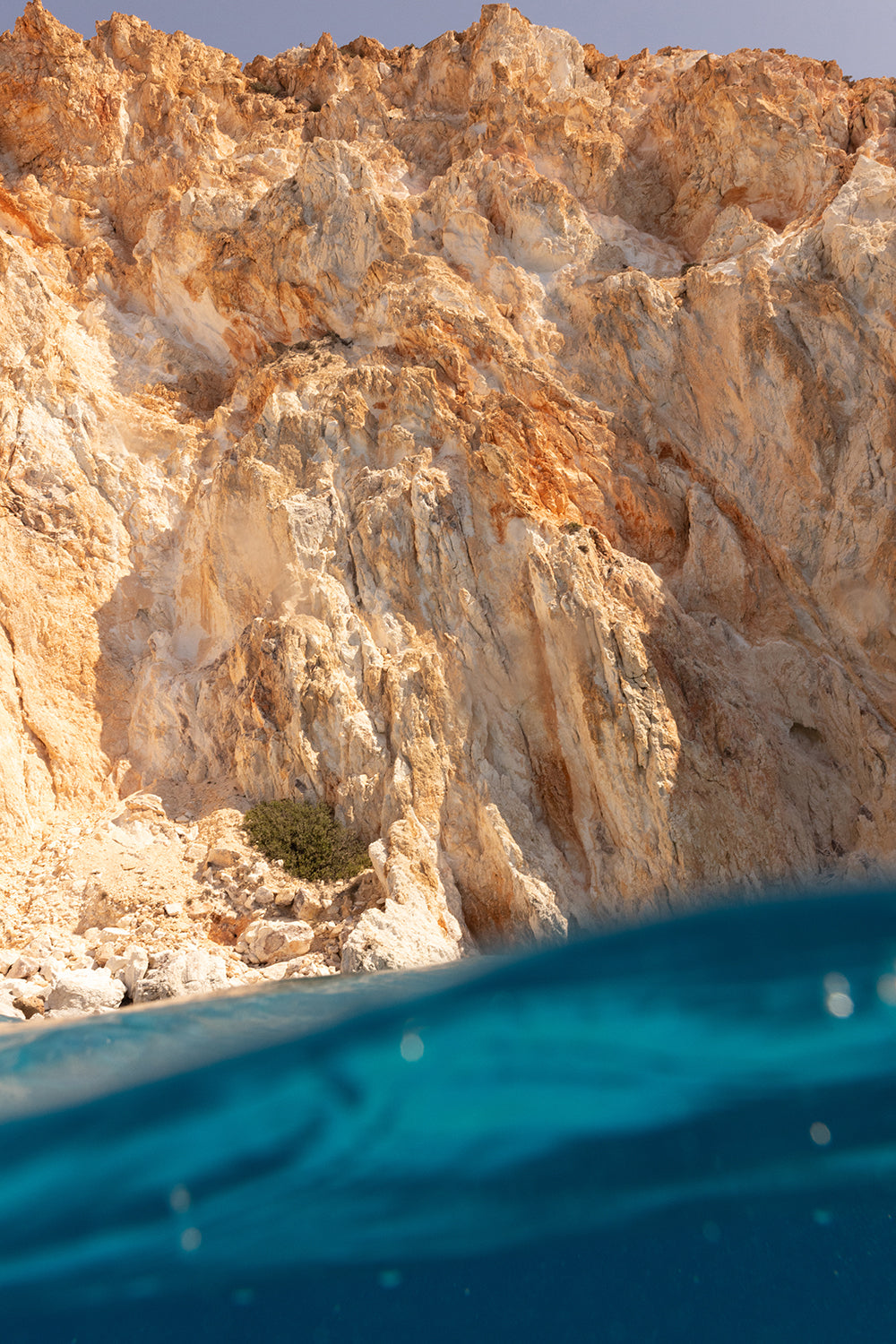 Captured with archival pigments on fine art paper, "Magic in Milos #7" by Francesca Owen showcases a rocky cliff rising majestically above the clear blue ocean. The rough, jagged texture of the cliff in warm brown tones offers a stunning contrast to the smooth surface of the water in the foreground.