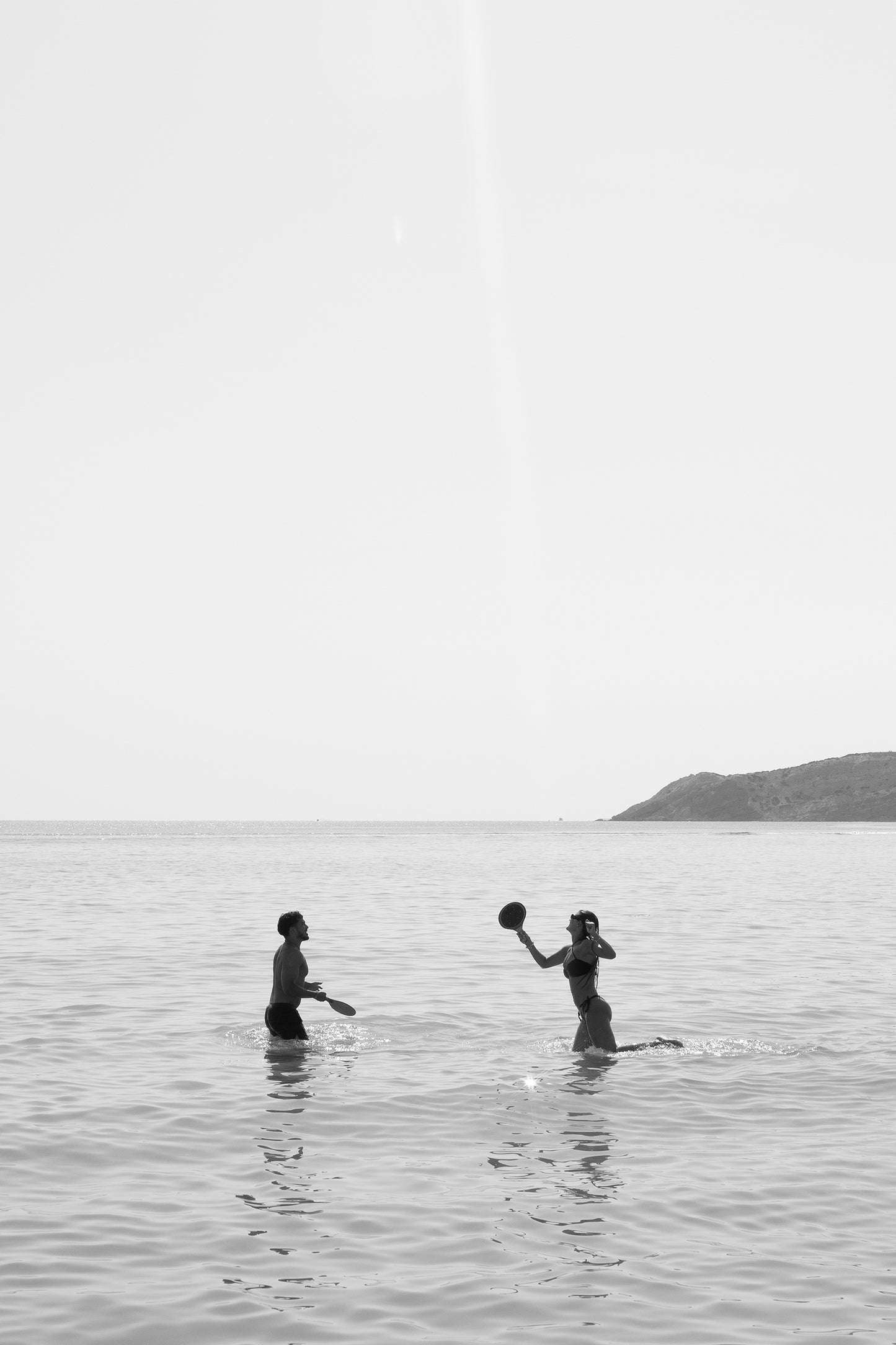 Title: "Magic in Milos #8" by Francesca Owen

Two people are engaging in a playful moment with a beach ball in shallow ocean waters, depicted in striking black and white. One is poised to catch or throw the ball, while the horizon and a distant hill frame the scene. This fine art print uses archival pigments on quality paper and includes a certificate of authenticity.