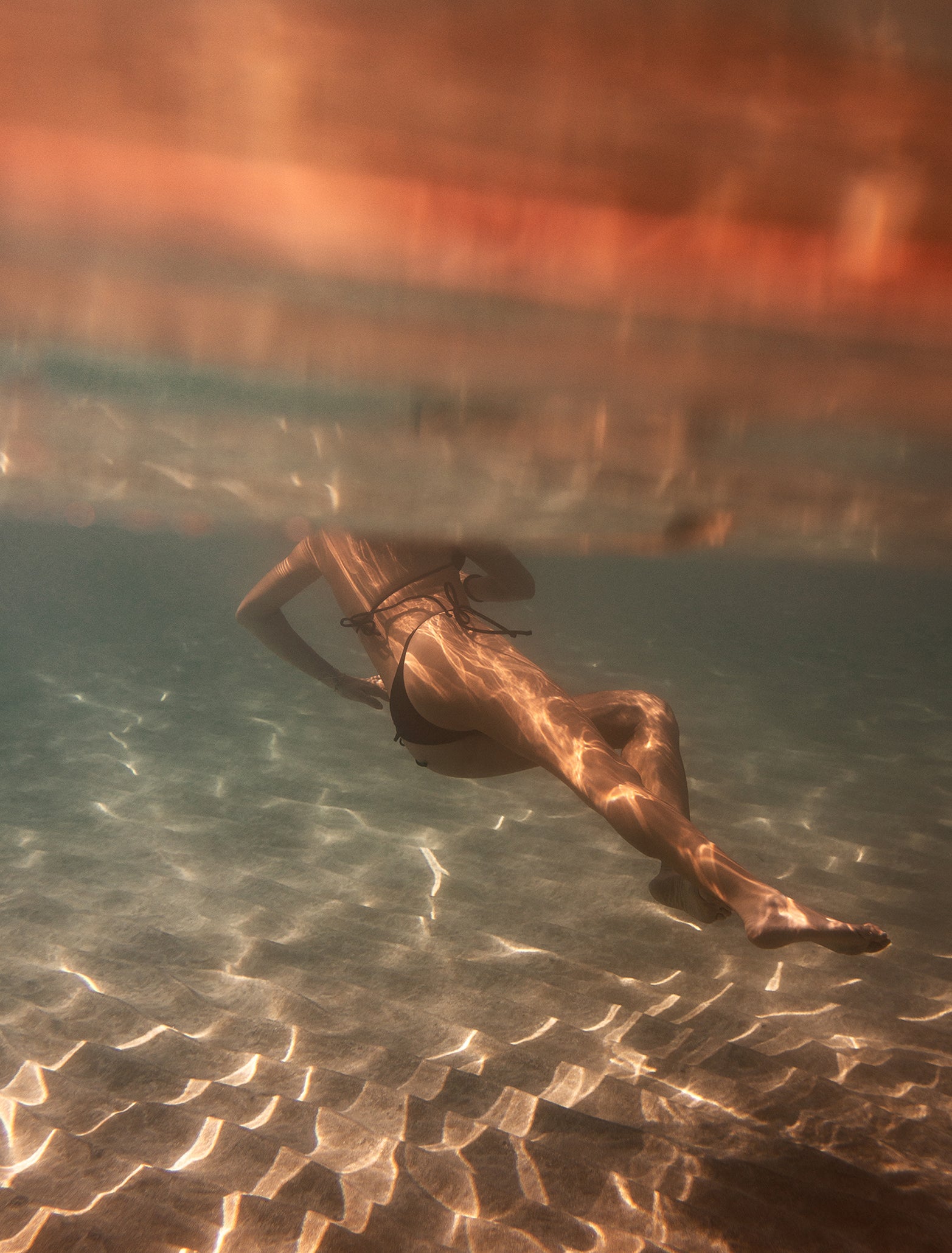 Swimming beneath the sunlit waters, a person clad in a swimsuit moves gracefully through the pool. The golden reflections create warm, wavy patterns of light that transform the scene into an artistic and serene atmosphere. In this depiction reminiscent of fine art photography, titled "Magic in Milos #3" by Francesca Owen, the swimmer's back elegantly glides through the rippling shadows.