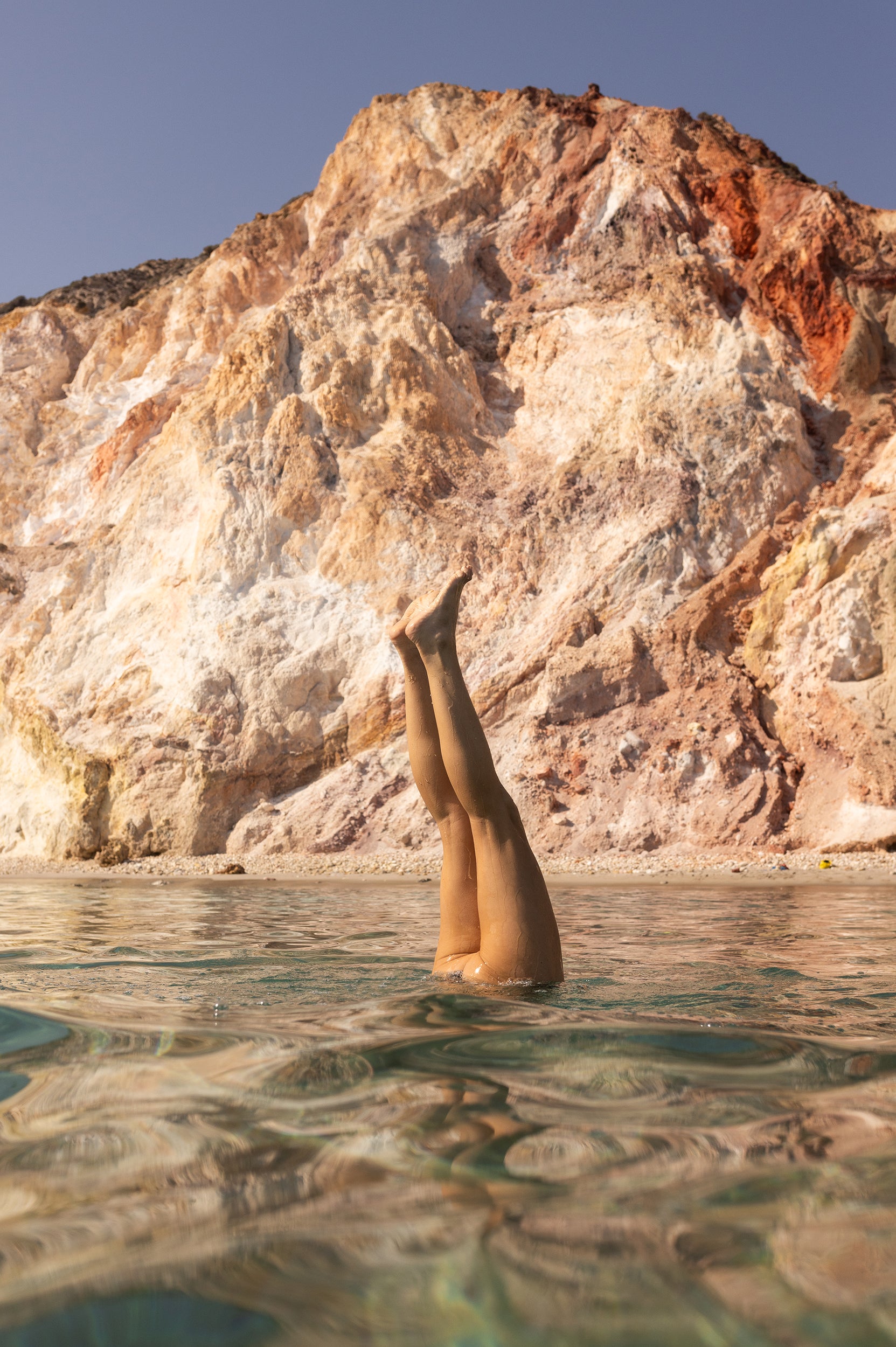 In "Magic in Milos #10" by Francesca Owen, a person is depicted doing a handstand in crystal-clear water with their legs extending above the surface, framed by a vibrant and rocky multicolored cliff under a clear sky. This striking image is produced using archival pigments on fine art paper and includes a certificate of authenticity.