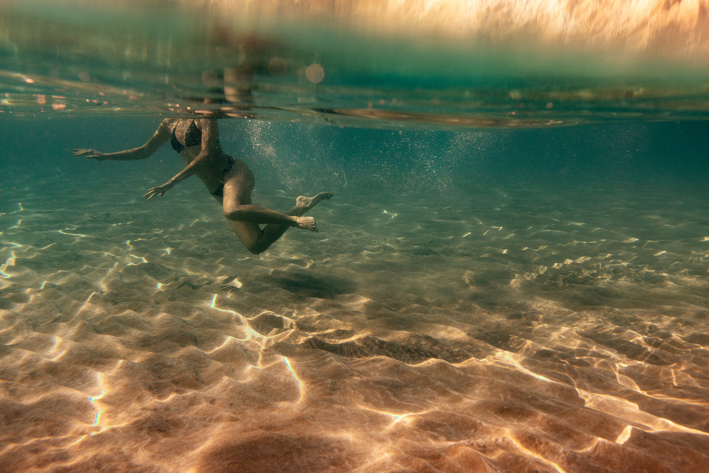 A person swims underwater in the crystalline waters of the ocean, with sunlight creating dancing patterns on the sandy sea floor. This peaceful and vivid scene, captured both above and below the surface, would make for a timeless display when printed on fine art paper using archival pigments from Francesca Owen's collection, "Magic in Milos #4.