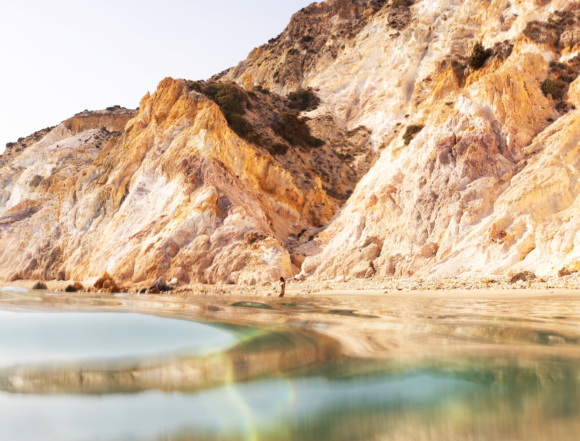 The "Magic in Milos #9" by Francesca Owen showcases a tranquil beach scene with captivating orange-tinted cliffs mirrored in the serene, clear water. A solitary figure walks along the shore beneath a clear sky, all meticulously captured on fine art paper with archival pigments to preserve each detail for generations.