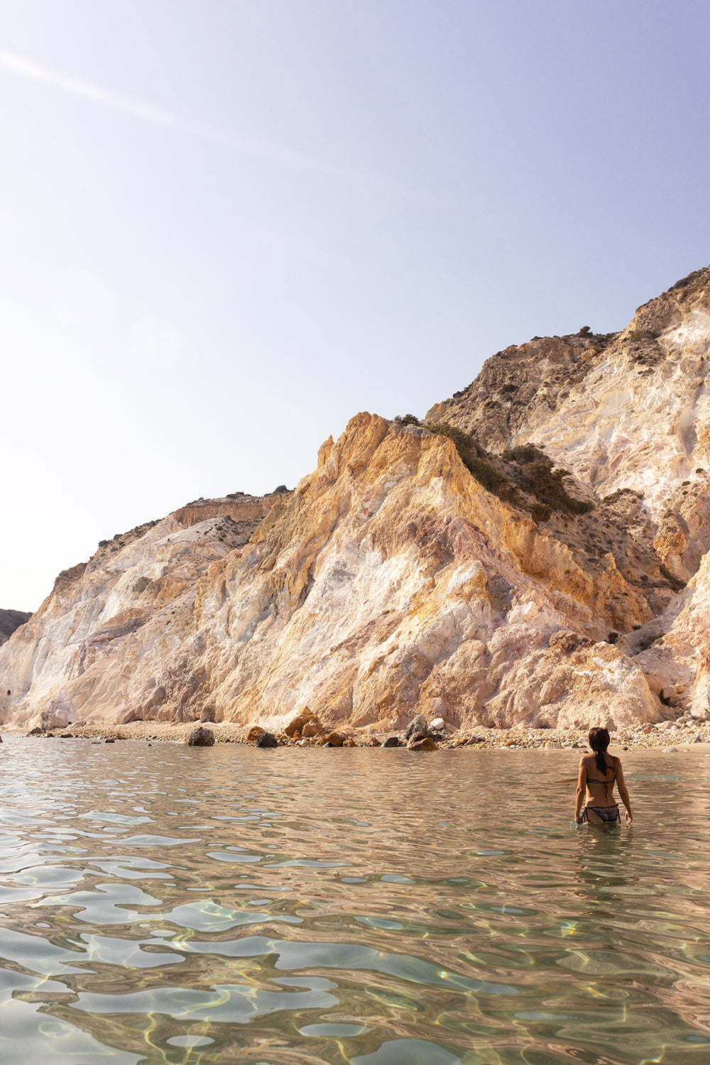 A person wades in the clear, calm waters of the sea near a rocky, sunlit cliff, with the scene basking in a warm glow. This exquisite moment is captured by Francesca Owen in "Magic in Milos #11" using archival pigments on fine art paper, each print accompanied by a certificate of authenticity.