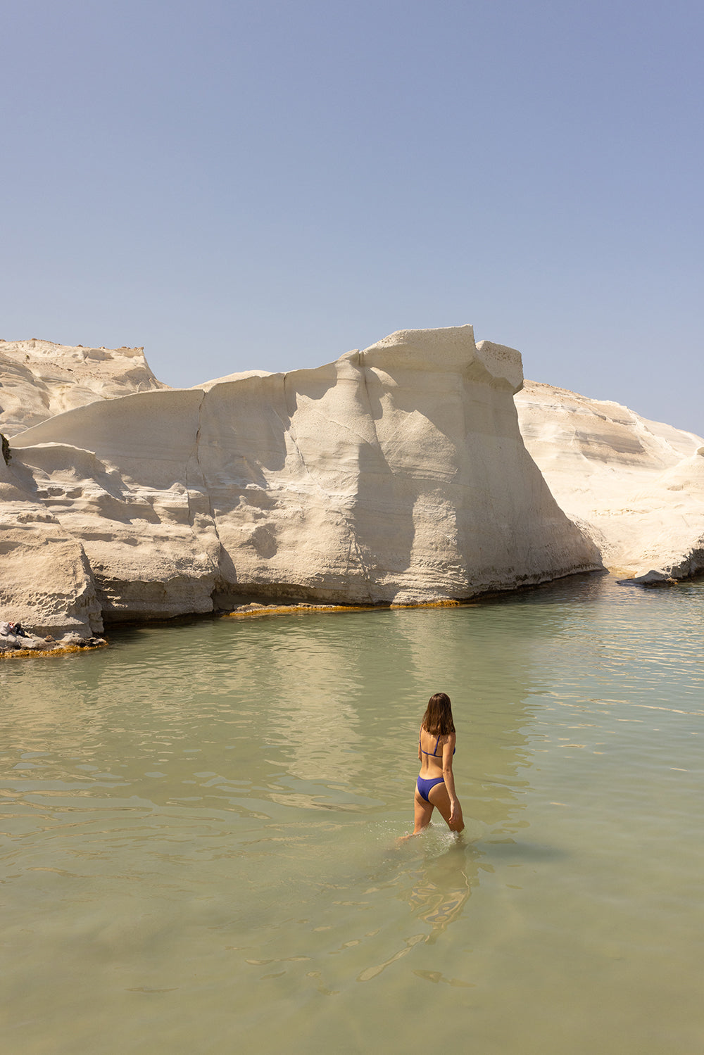 A person wearing a blue swimsuit walks through shallow water towards large white rock formations under a clear blue sky, beautifully depicted with archival pigments on fine art paper for long-lasting vibrancy. This artwork, titled "Magic in Milos #12," is created by Francesca Owen and comes with a certificate of authenticity.