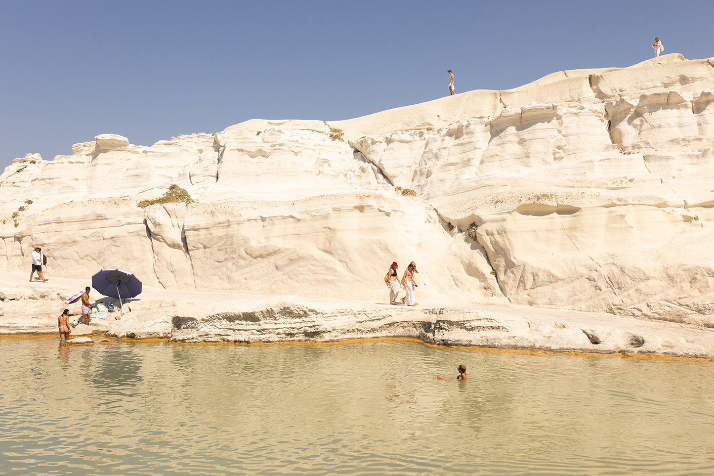 Visitors relish a sunny day on the beach, surrounded by striking white cliffs. A few people take leisurely strolls, while a swimmer enjoys the tranquil water. An umbrella offers shade to some along the shore as they bask under the clear blue sky, creating a picturesque scene reminiscent of "Magic in Milos #13" by Francesca Owen—a piece captured on fine art paper with archival pigments for enduring beauty.