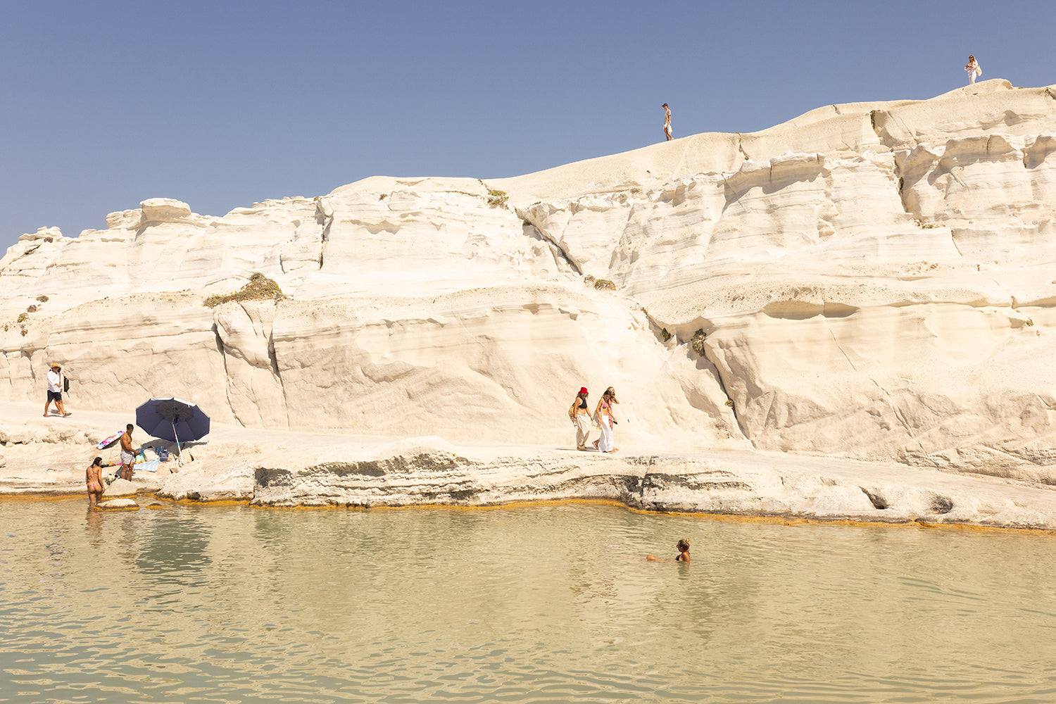 Visitors relish a sunny day on the beach, surrounded by striking white cliffs. A few people take leisurely strolls, while a swimmer enjoys the tranquil water. An umbrella offers shade to some along the shore as they bask under the clear blue sky, creating a picturesque scene reminiscent of "Magic in Milos #13" by Francesca Owen—a piece captured on fine art paper with archival pigments for enduring beauty.