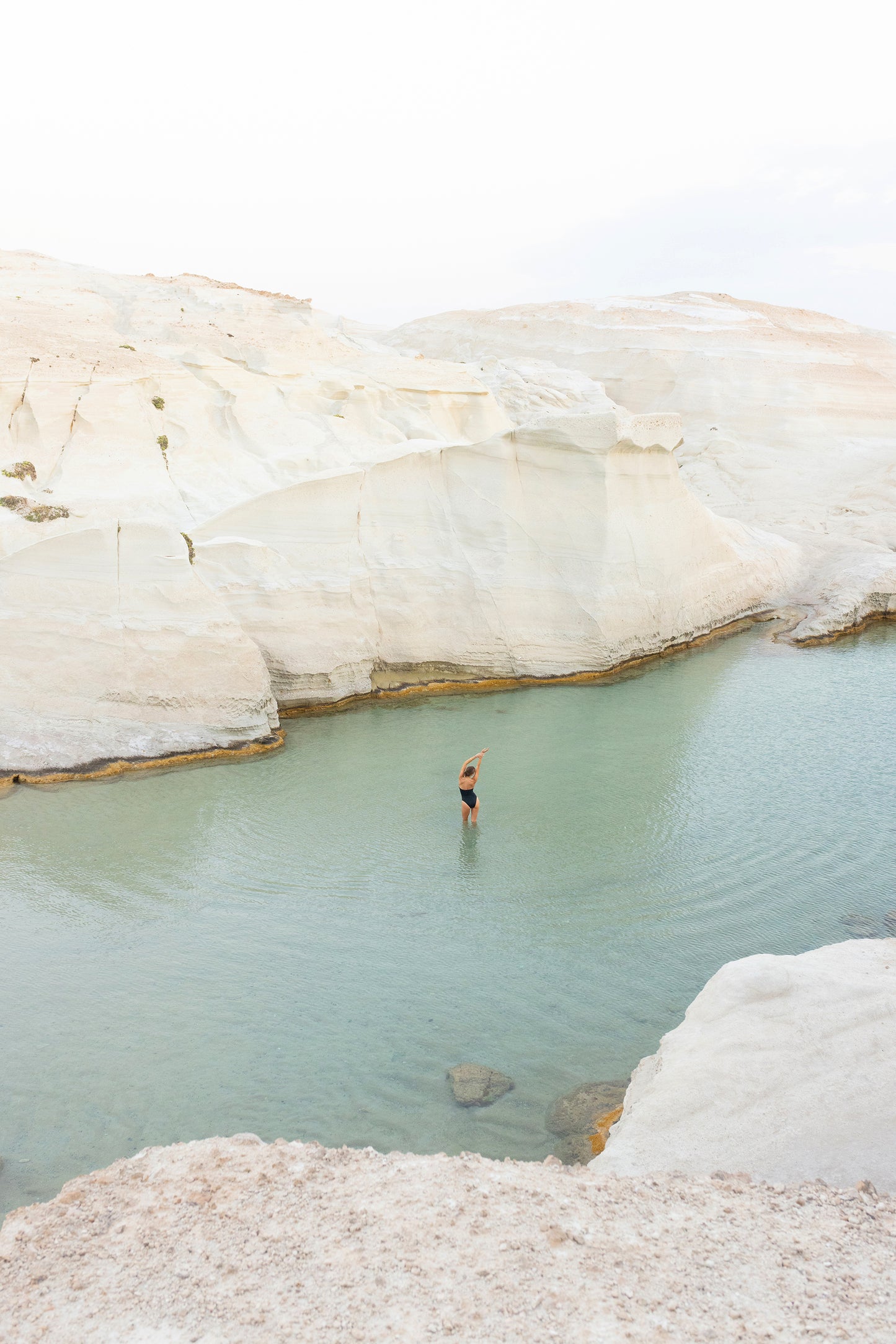 A person stands in a clear, shallow pool surrounded by white, rocky cliffs under a pale sky. This serene landscape is evocative of the tranquil beauty captured in Francesca Owen's artwork, Magic in Milos #2, framed by natural rock formations and calm water.