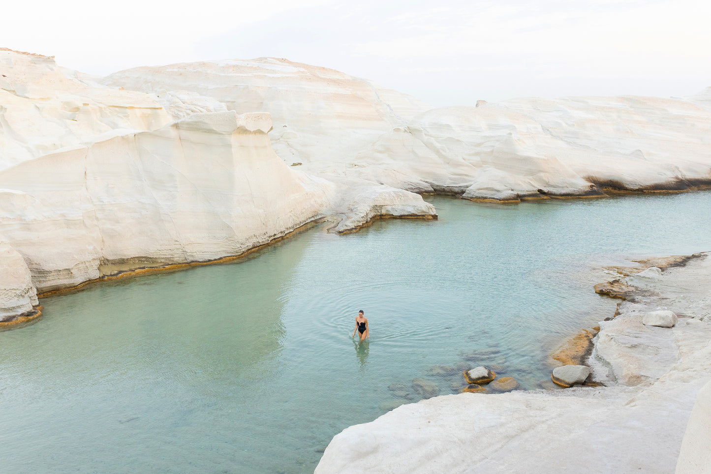 A person stands in a calm, shallow pool of clear water surrounded by large, smooth, white rock formations under a cloudy sky. The serene and tranquil atmosphere is captured perfectly in Francesca Owen's *Magic in Milos #1*, printed on fine art paper with archival pigments.