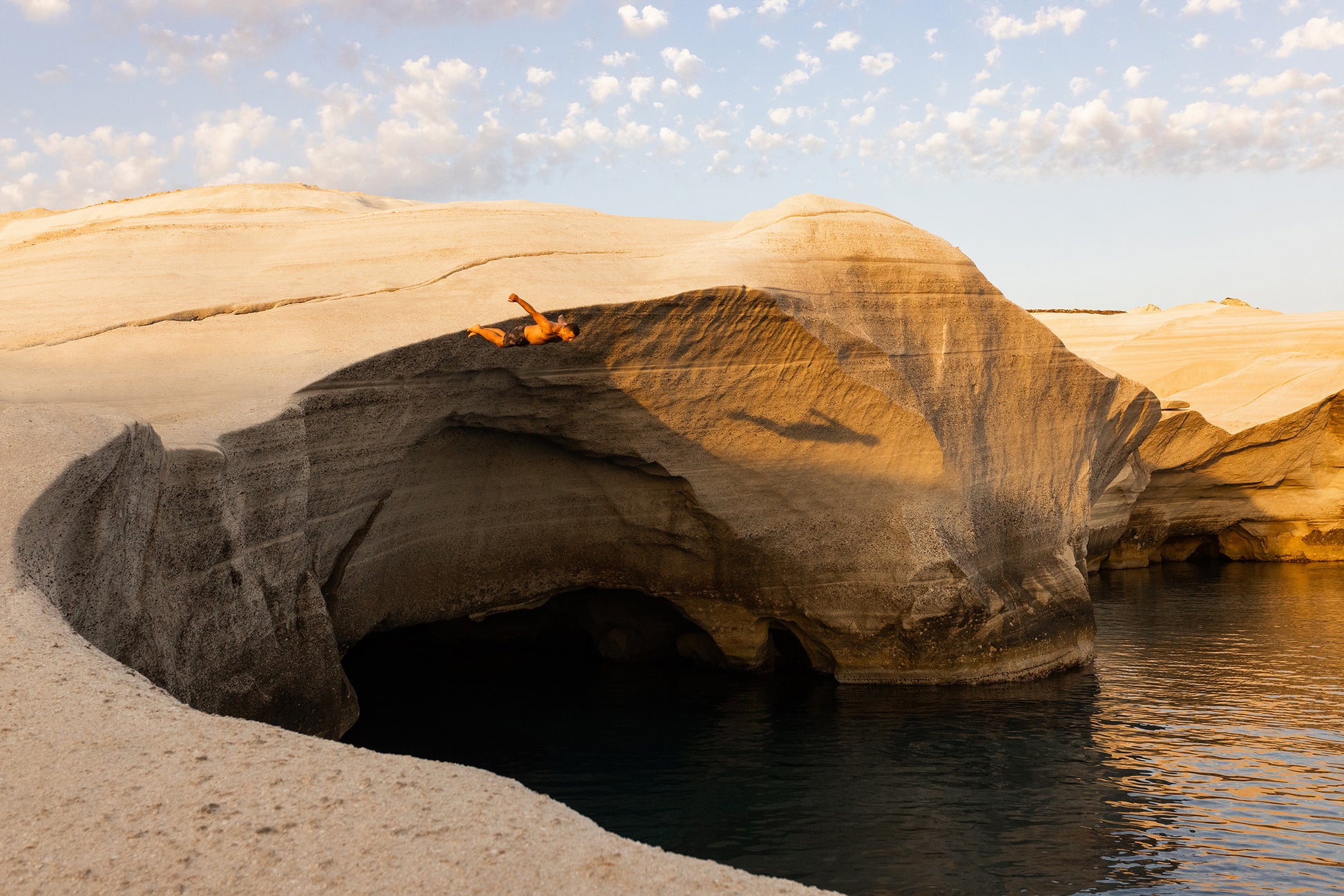 Basking on the sunlit, white rock formation of "Magic in Milos #6" by Francesca Owen, a person lies above a cave-like structure next to calm blue water. Scattered clouds fill the sky, casting warm, golden light over the scene, embodying nature itself as if captured on fine art paper.