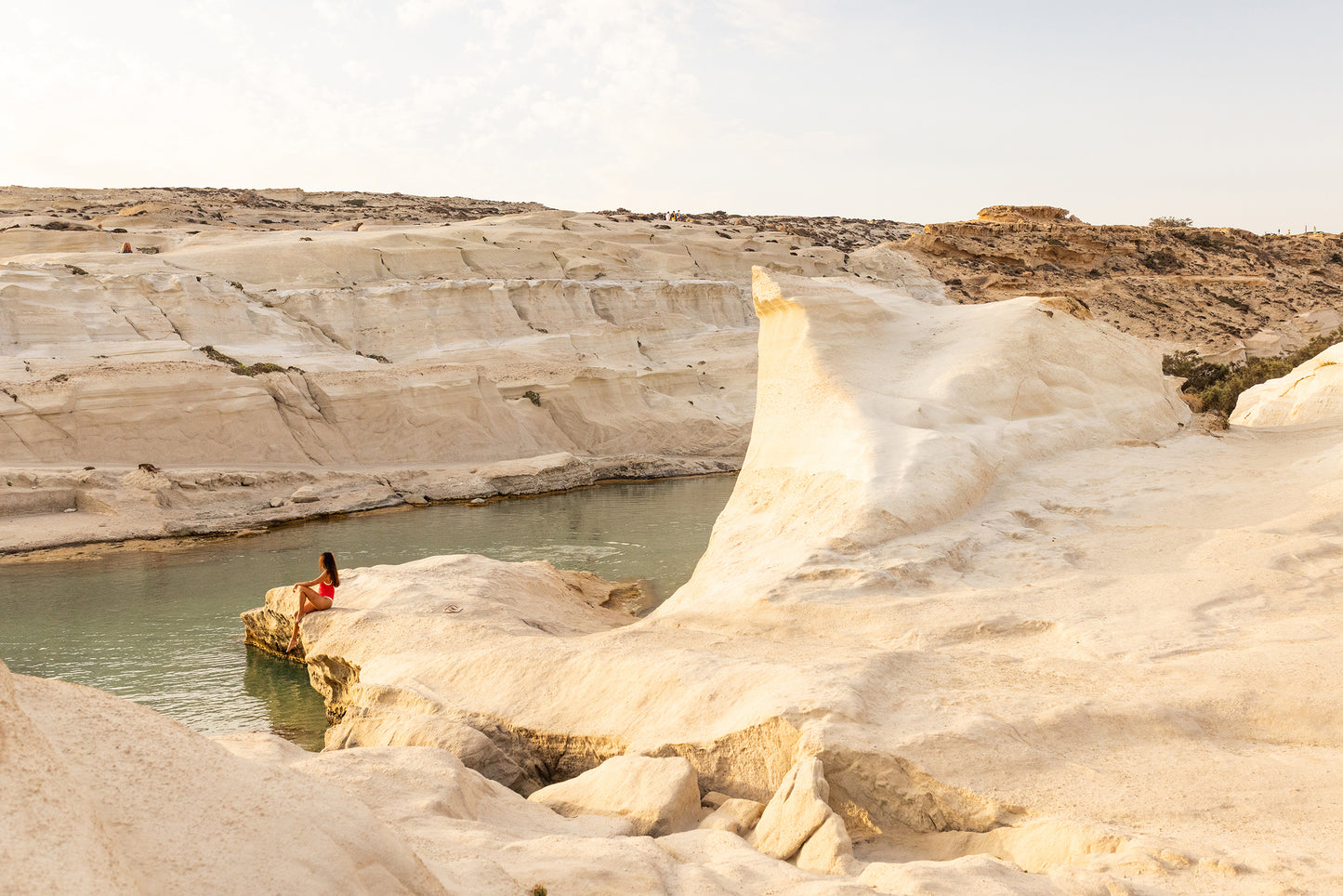 The "Magic in Milos #5" by Francesca Owen features a person in a red outfit sitting on a rock beside a tranquil water body, with dramatic white rock formations and clear skies, captured using archival pigments that evoke the quality of fine art paper.