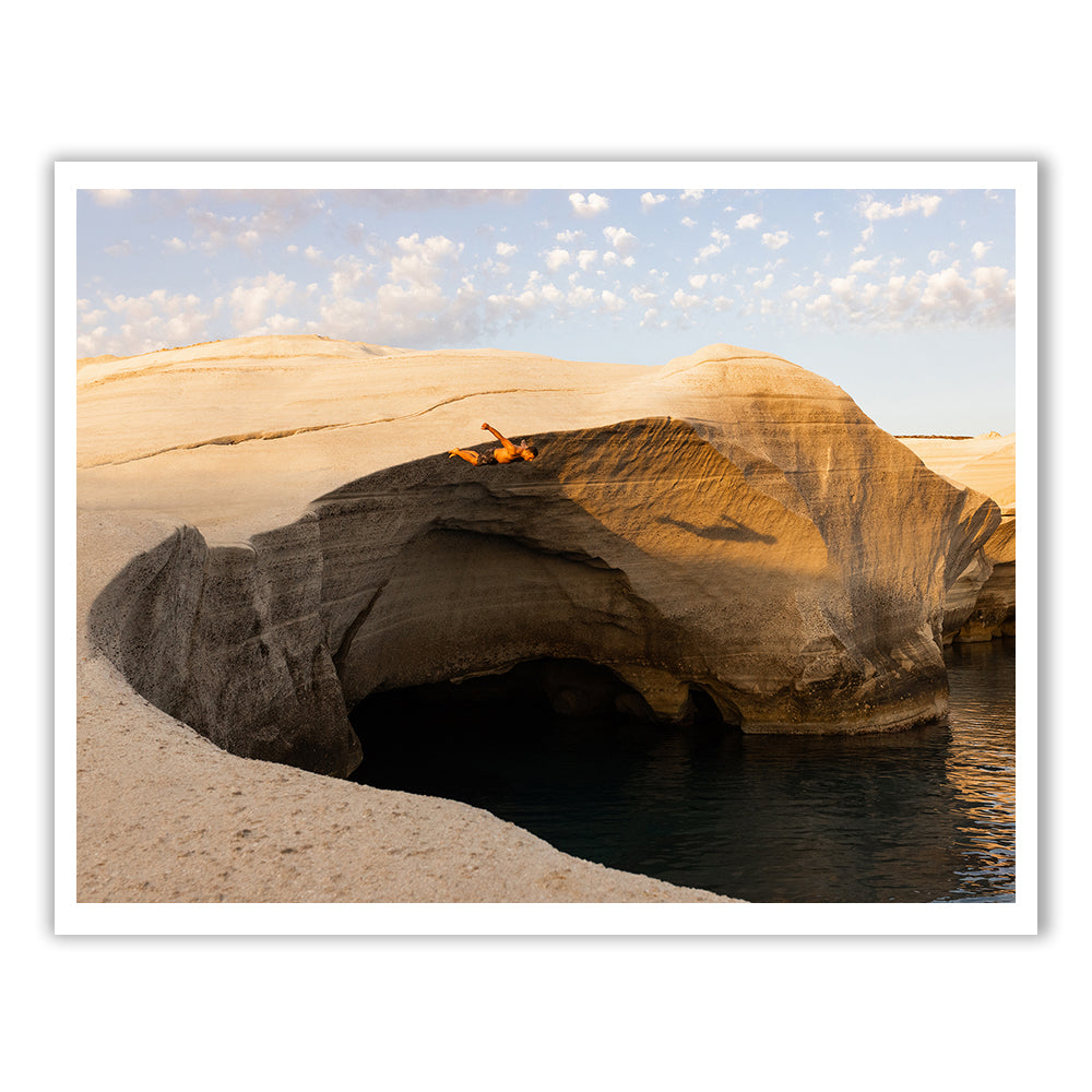 A person relaxes on large, smooth, white rock formations overlooking a deep blue sea, where the partly cloudy sky enhances the scene's tranquility—a moment as serene as "Magic in Milos #6" by Francesca Owen, crafted with archival pigments on fine art paper.