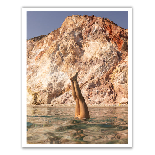 A person performs a handstand underwater in the clear ocean, their legs breaking the surface. In the background, a large rocky cliff showcases shades of brown and orange under a blue sky. Captured with archival pigments on fine art paper, this moment is embodied in "Magic in Milos #10" by Francesca Owen.