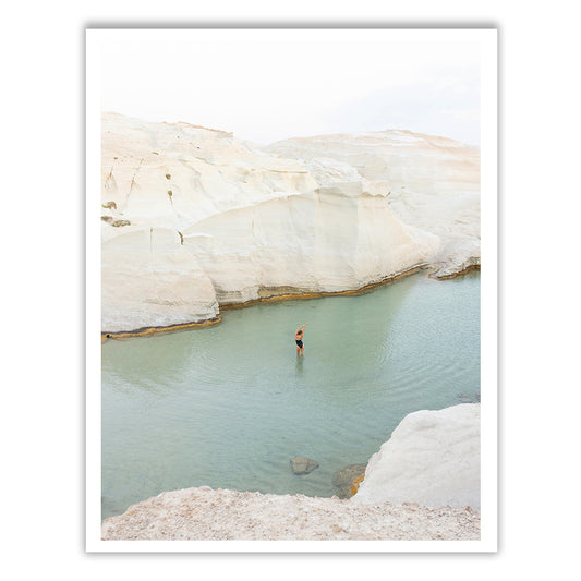 A person stands in a shallow, greenish-blue body of water surrounded by white rocky cliffs, their arm raised in a graceful gesture. This tranquil scene evokes the timeless beauty of nature, reminiscent of the serene landscapes captured by Francesca Owen's "Magic in Milos #2.