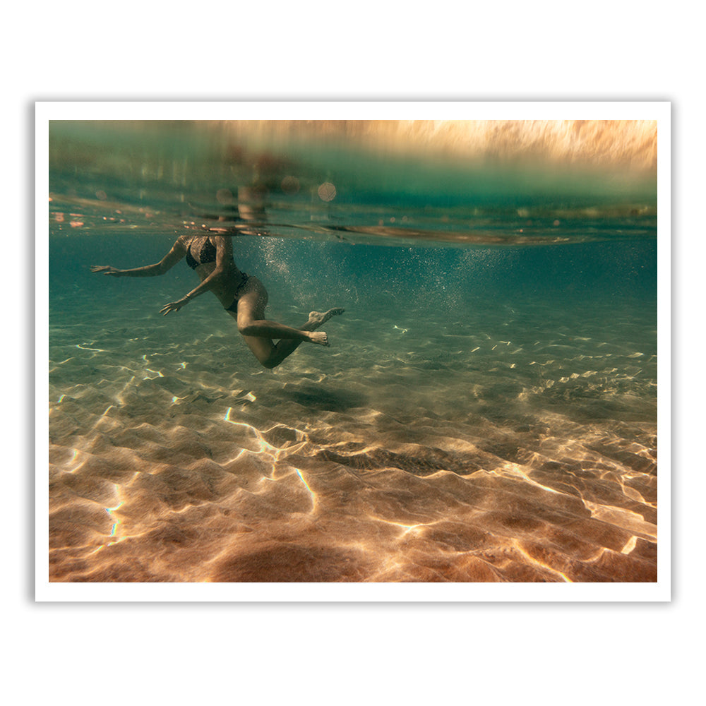 In "Magic in Milos #4" by Francesca Owen, a person swims gracefully underwater in the clear ocean, with golden sunlight illuminating the sandy seabed. This tranquil and liberating scene is beautifully captured using archival pigments on fine art paper and comes with a certificate of authenticity.