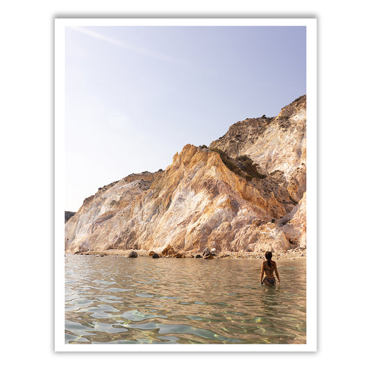 A person stands in clear, shallow water near a rocky shoreline. Sunlit cliffs rise majestically in the background under a clear sky, evocative of the exquisite scene captured in Francesca Owen's "Magic in Milos #11," worthy of archival pigments on fine art paper.