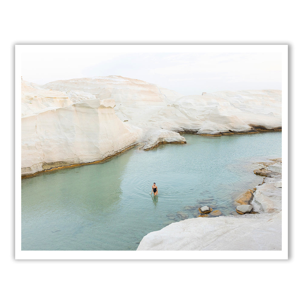 A person is immersed in a tranquil, turquoise water pool encircled by smooth, white rock formations beneath a gentle sky. This serene and pristine landscape resembles "Magic in Milos #1," an exquisite artwork by Francesca Owen, printed on fine art paper with archival pigments to preserve its timeless beauty.