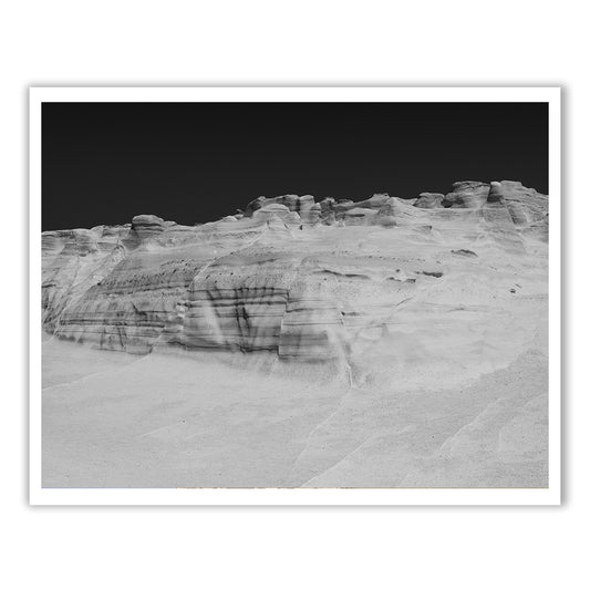 Introducing the "Magic in Milos #14" by Francesca Owen—a black and white photograph depicting a barren desert landscape with eroded rock formations under a dramatic sky. This stunning piece is crafted using archival pigments on fine art paper, ensuring longevity and depth of contrast. It comes with a certificate of authenticity.