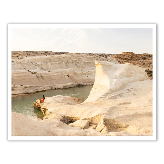 The serene landscape of "Magic in Milos #5" by Francesca Owen beautifully captures a person dressed in red, sitting on a rocky ledge by tranquil water. This scene is framed by smooth, sand-colored formations and set beneath a partly cloudy sky, all expertly rendered using archival pigments.