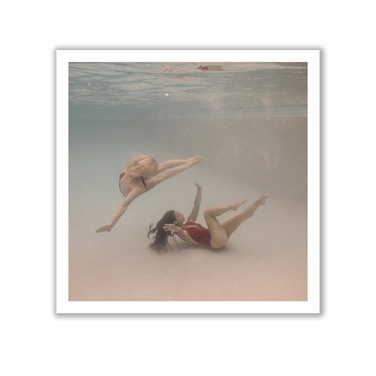 Two women are underwater, clad in swimsuits. One woman, with light-colored hair, gracefully swims above the other, who has dark hair. They are enveloped by crystal-clear water, exuding fluidity and tranquility. Titled "Opposites Attract #2," this artwork by Francesca Owen is captured on fine art paper with archival pigments and comes with a certificate of authenticity.