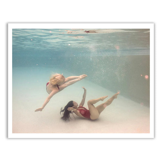 Two individuals in swimsuits gracefully execute a synchronized underwater dance in a pool, reminiscent of fine art paper. One person dives forward with arms extended, while the other sits reaching upward. Bubbles and soft lighting enhance the dreamlike scene, capturing Francesca Owen's signature style in "Opposites Attract 02.